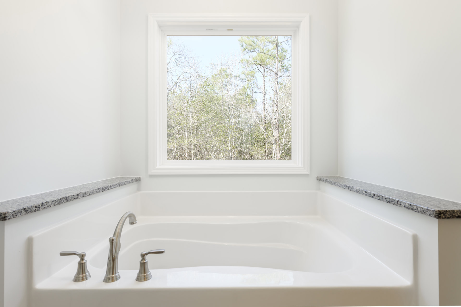 Freestanding white bathtub beneath a large window with tree views, chrome faucet, and light-colored bathroom walls