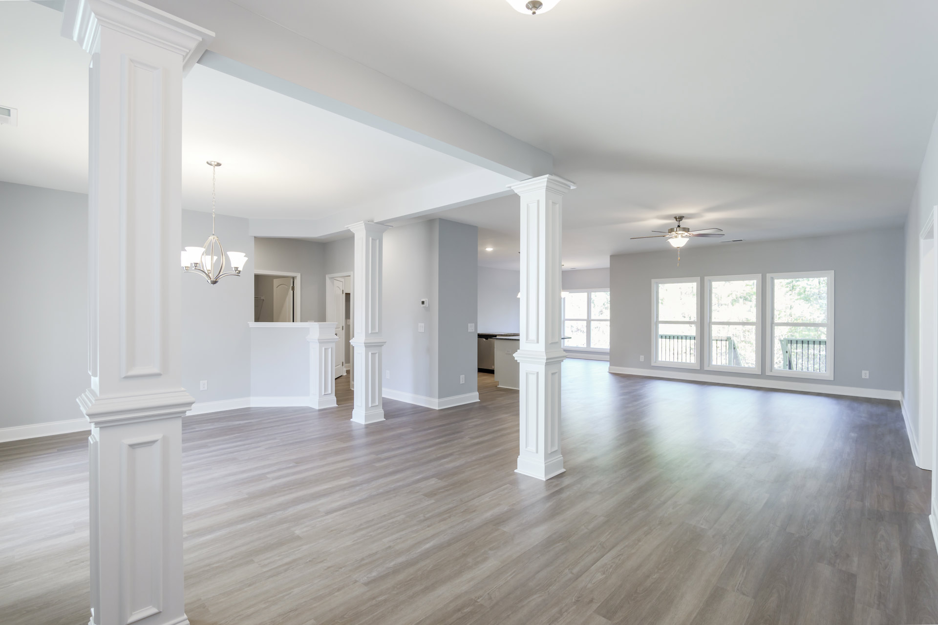 Spacious living area with polished hardwood floor, white pillars, chandelier, large window overlooking trees, and recessed ceiling lights