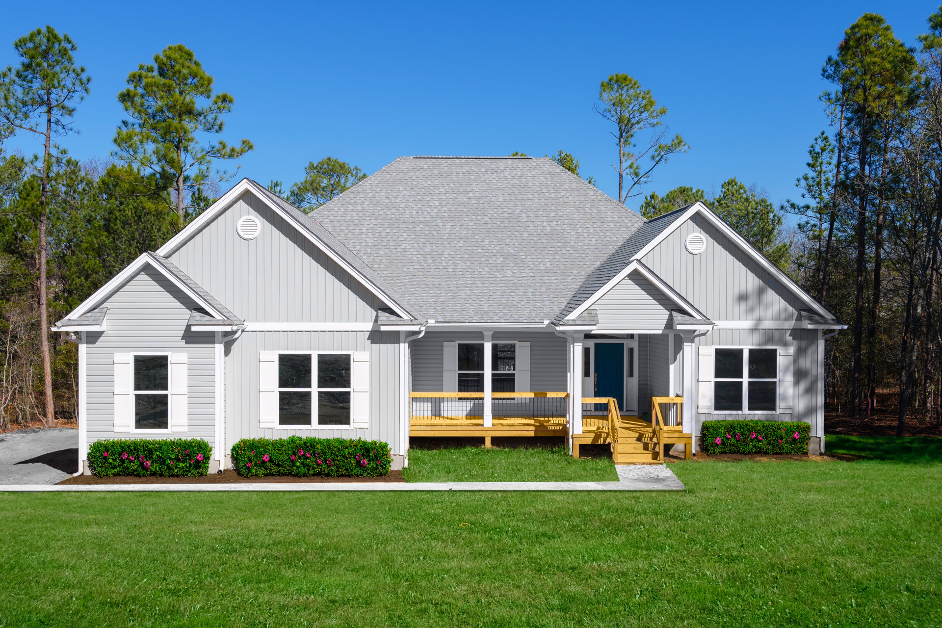 Two-story house with white siding, black metal porch railing, wooden deck, manicured lawn, mature trees, and bushes with purple flowers in the yard