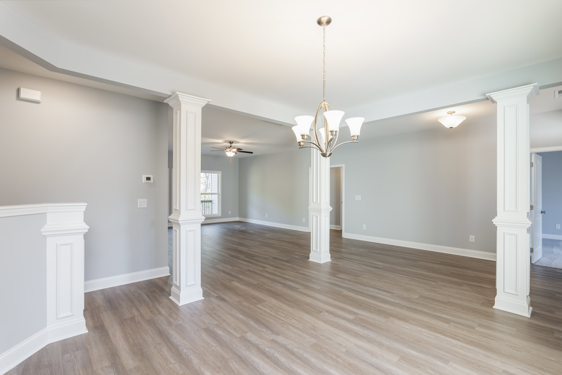 Spacious living area with wood flooring, white columns, ceiling fan, and large window overlooking trees