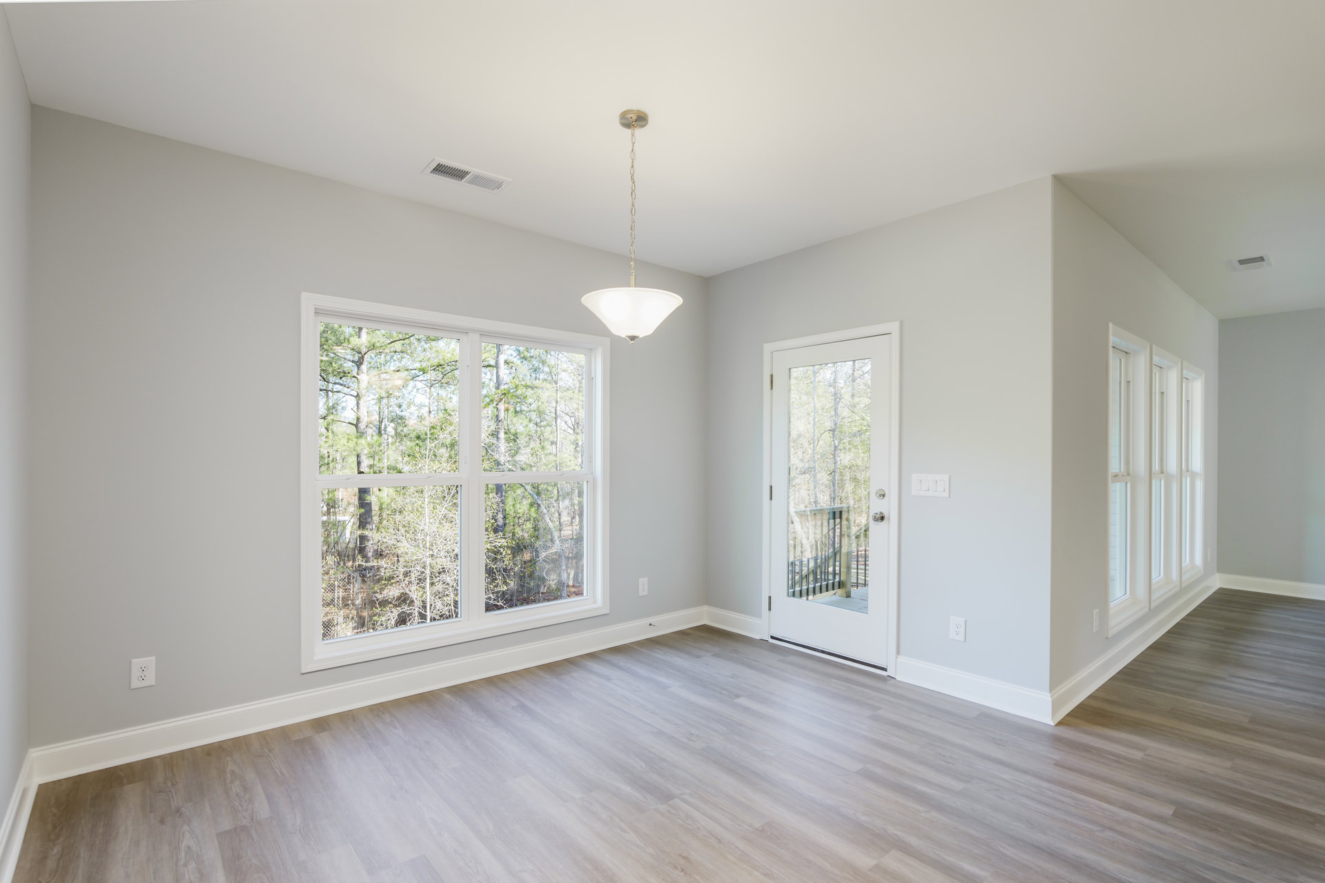 Bright room with wood flooring, white plaster walls, large window showing trees outside, ceiling light fixture, and door with outdoor view.