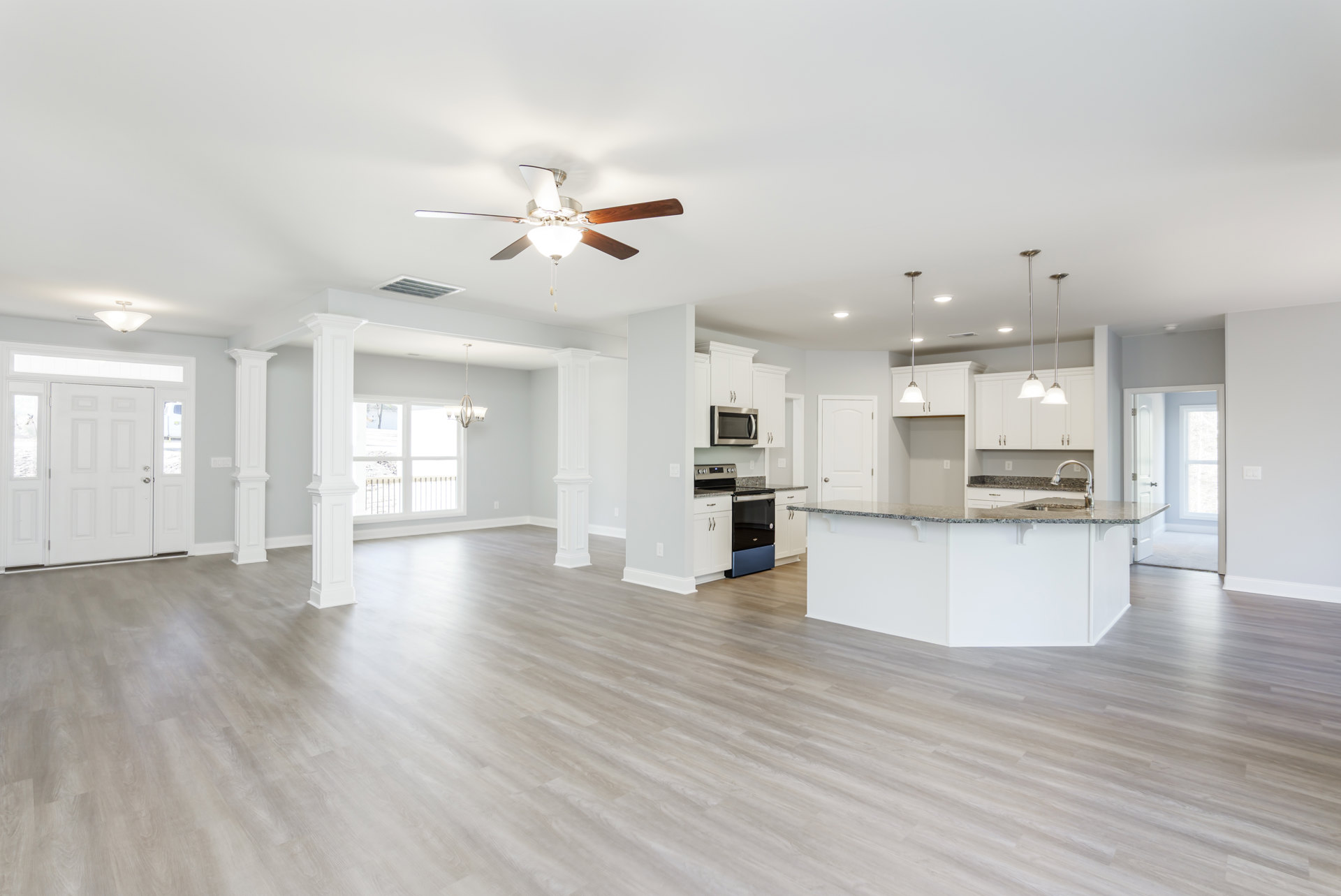 Spacious open floor plan featuring a kitchen with wood laminate flooring, white cabinetry, built-in microwave, adjacent dining area, ceiling fan, white column, and a white door