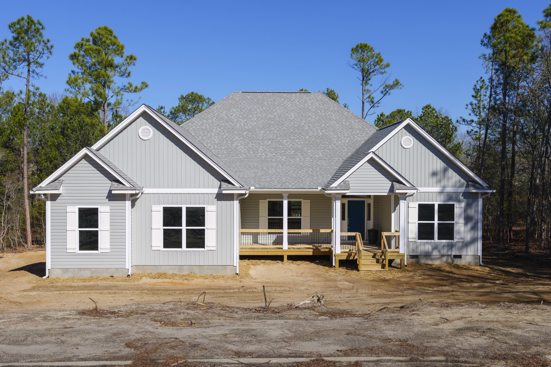 Partially built house with exposed wooden framing, white porch posts, unfinished wooden deck, black and white window frames, dirt driveway scattered with sticks, surrounded by