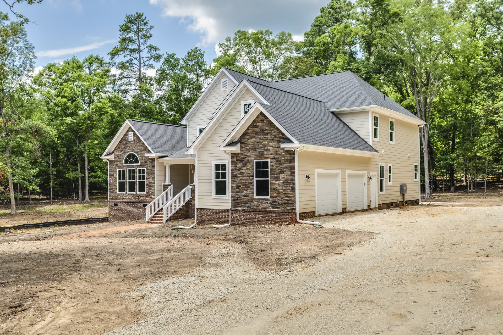 Two-story house with white-framed windows, brick and stone exterior walls, attached white garage door, concrete driveway, outdoor stairs, and mature trees surrounding the property