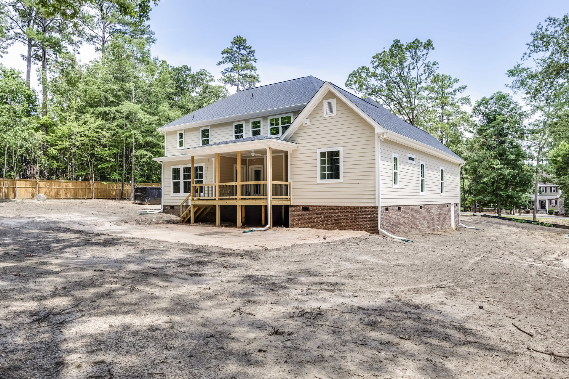 Two-story house under construction with multi-pane windows, wooden porch, metal railings, dirt yard, and trees in the background