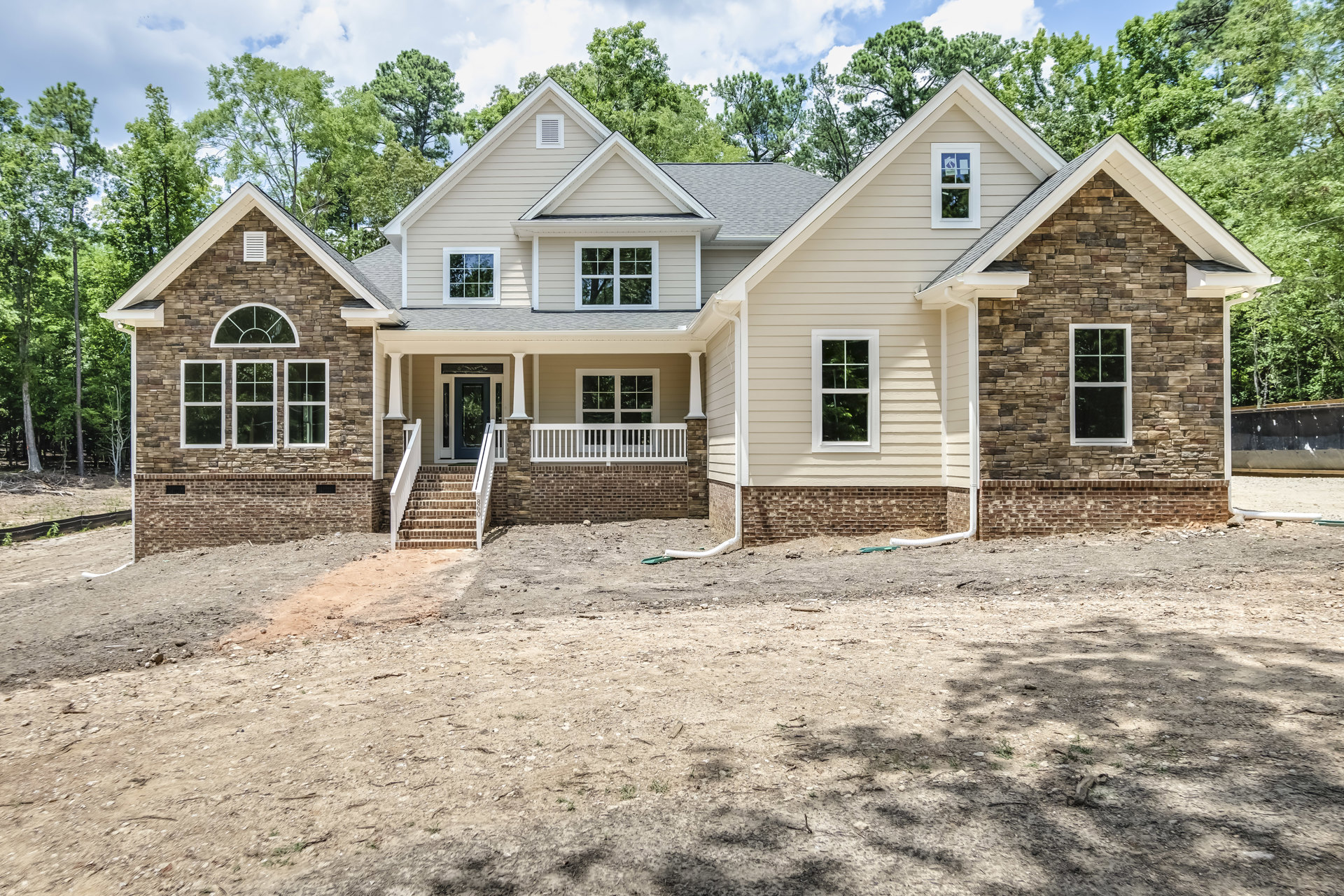 Two-story custom home with stone and brick exterior, large windows, covered porch, brick steps, concrete driveway, and mature trees in front yard