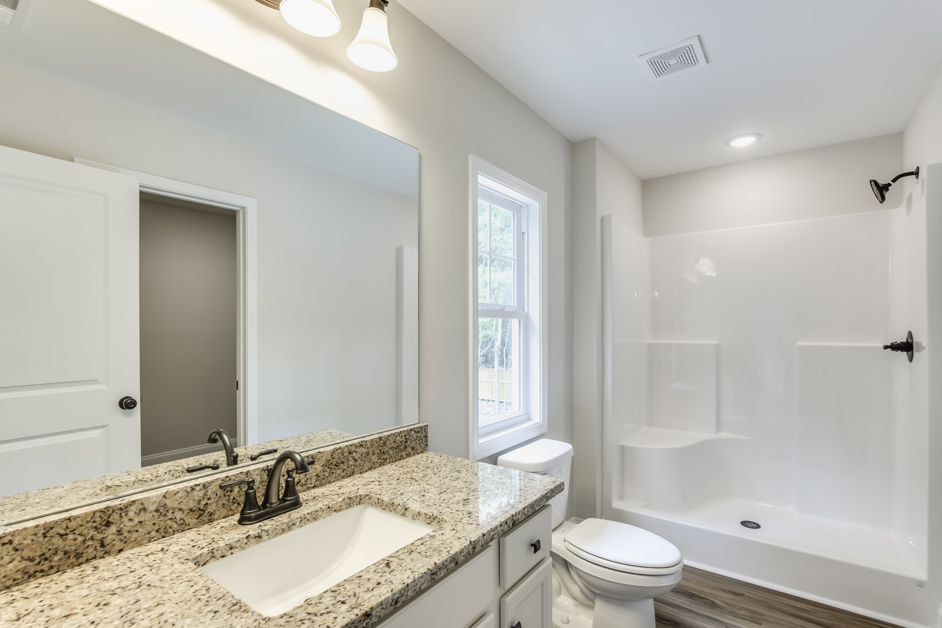 Bathroom with marble countertop sink, chrome faucet, glass-enclosed shower, white toilet, wall mirror, and window overlooking trees