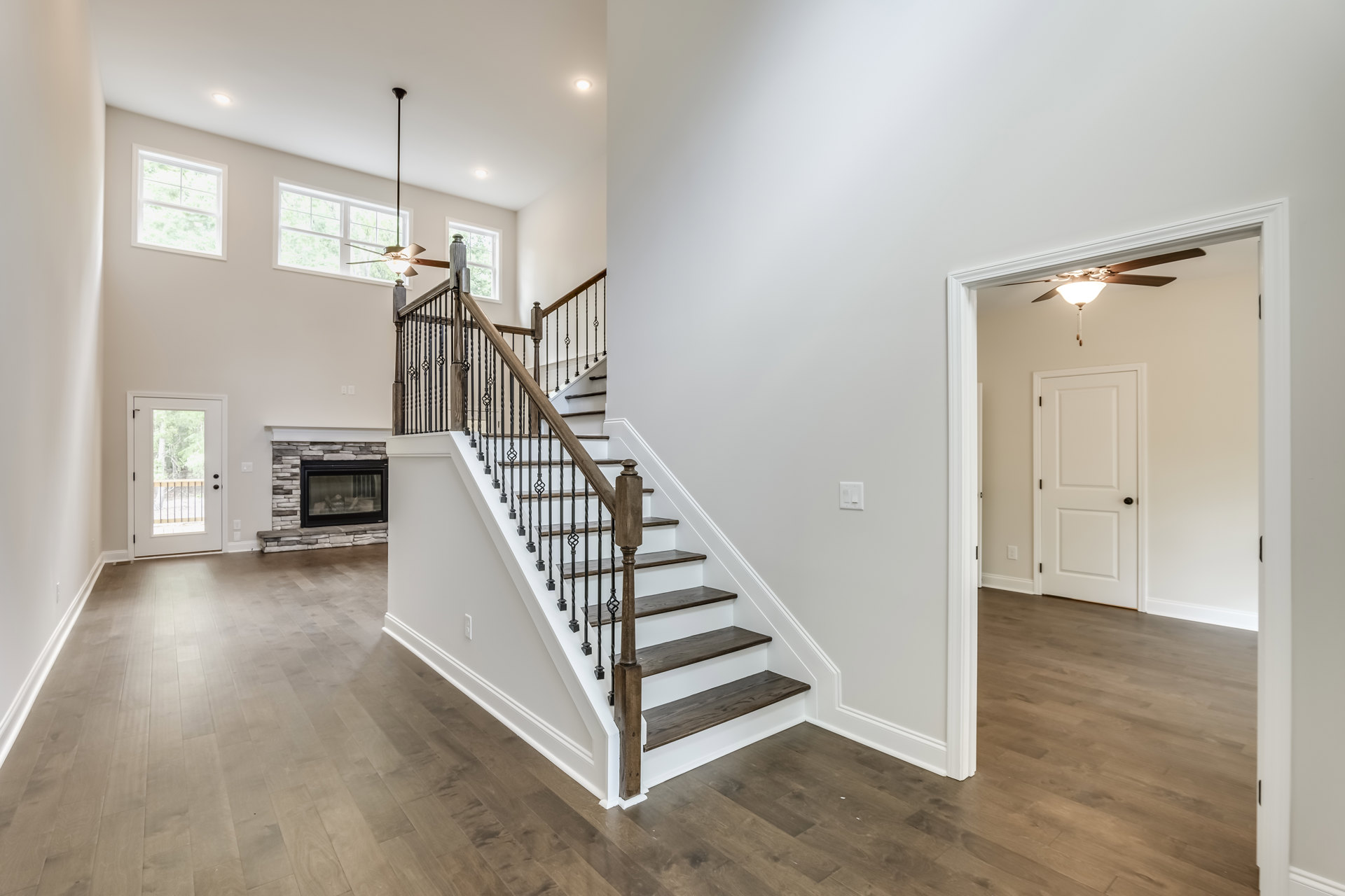 Wood staircase with matching handrail, white walls, glass window above fireplace, white door with black knob, laminate wood flooring, white-framed window, plaster ceiling.
