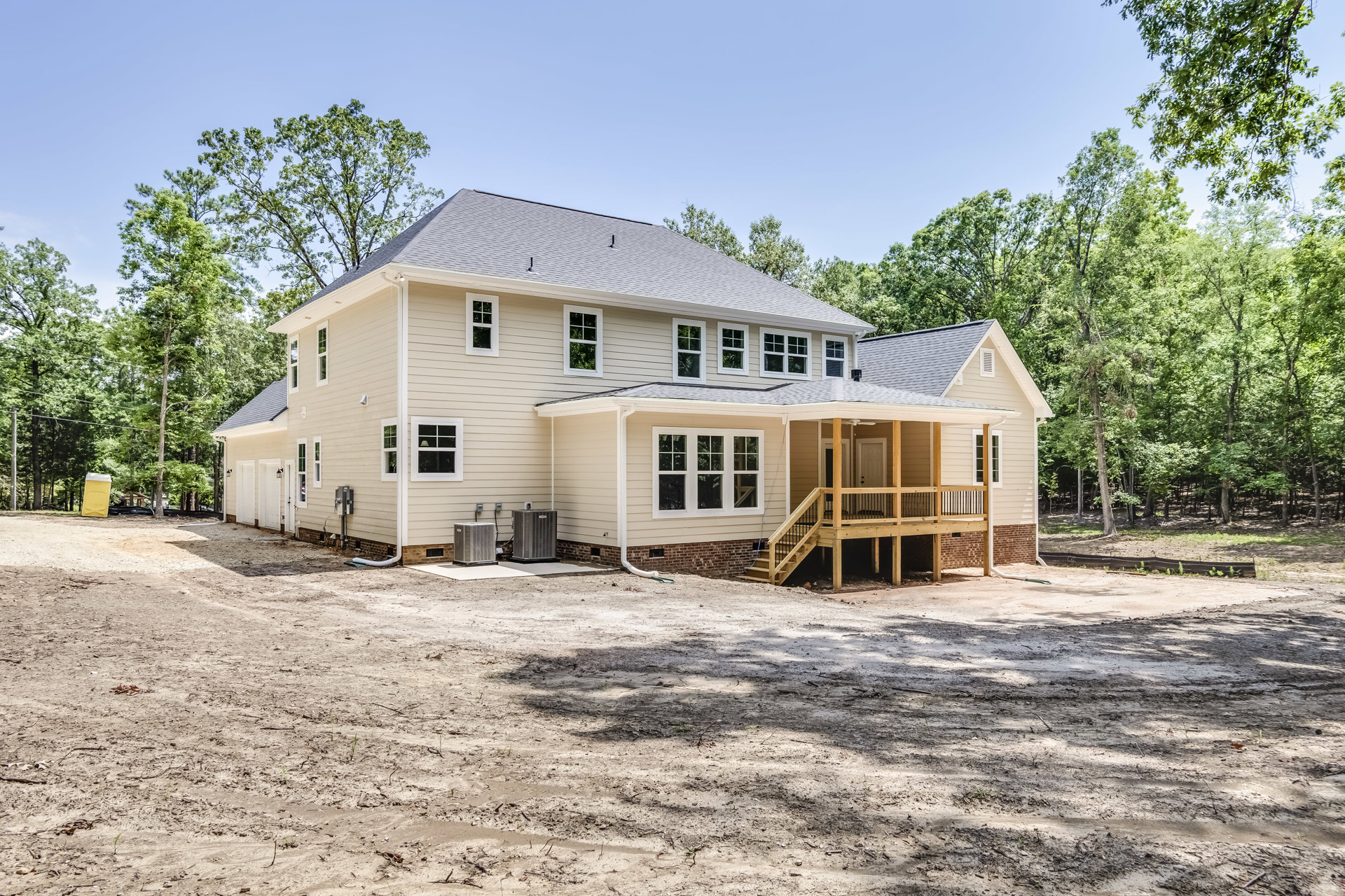 Two-story house with gray siding, wooden porch and railing, large gray heat pump on platform, dirt yard surrounded by mature trees, close-up window visible.