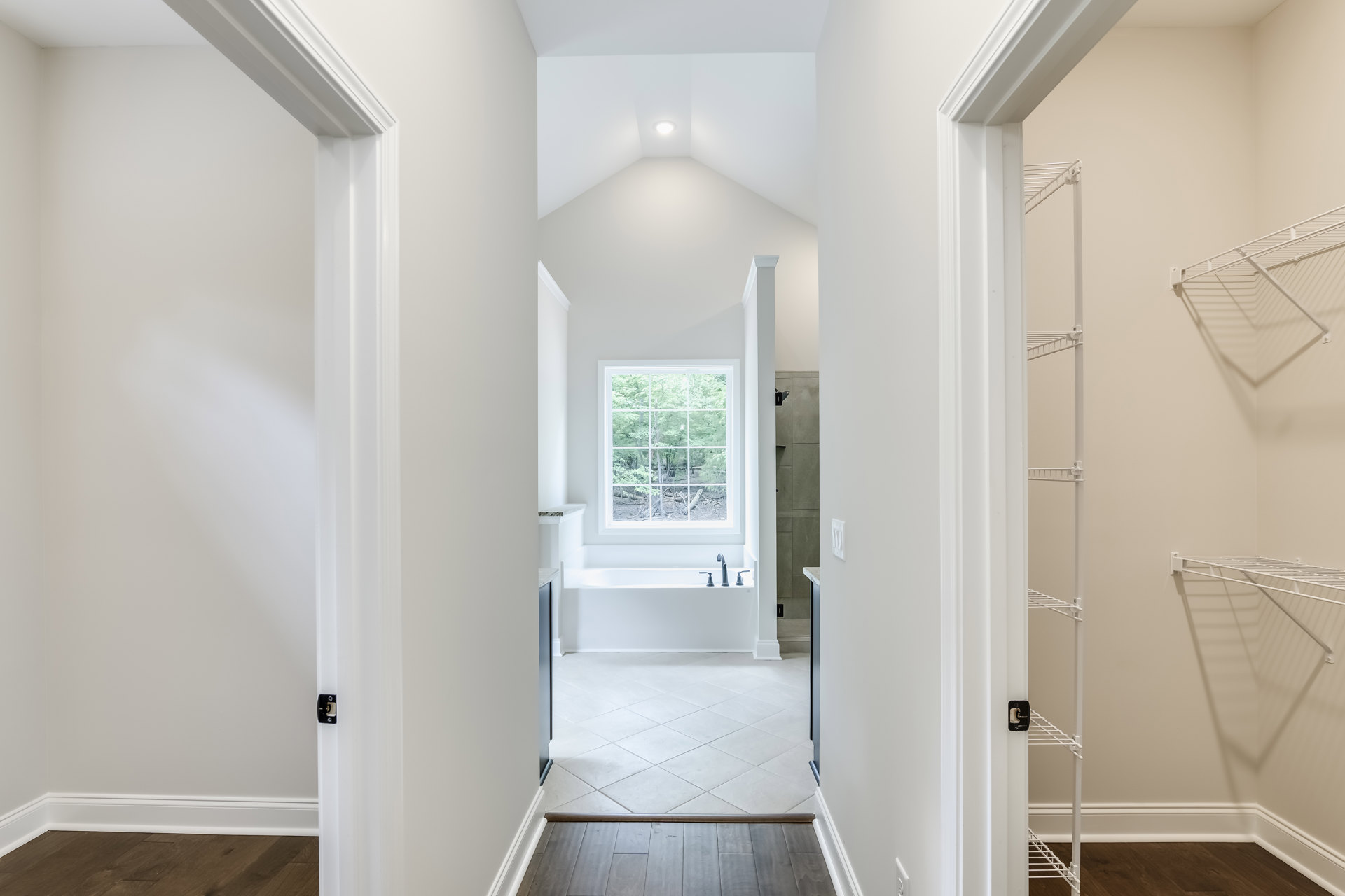 Hallway with white tile floor, white walls, built-in white shelf, large window overlooking trees, freestanding tub near window, wood flooring detail