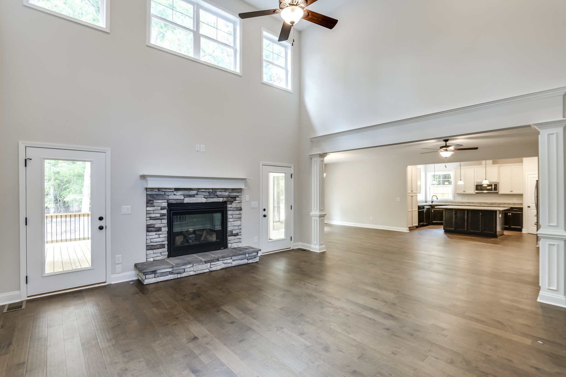 Spacious living room featuring wood flooring, central fireplace with glass doors, ceiling fan with light fixture, white door with window, and glass door opening to deck overlooking