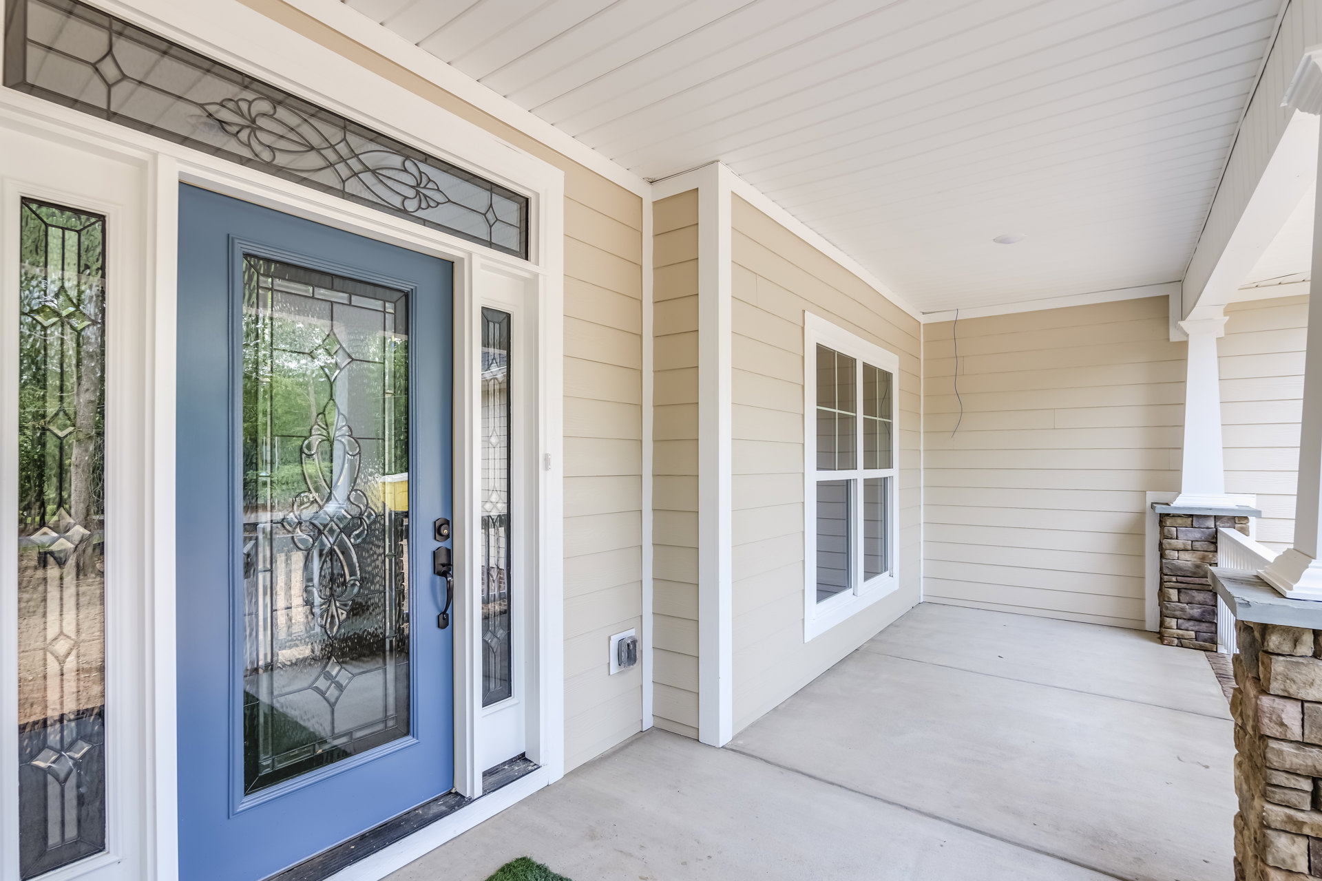 Blue front door with stained glass panel, covered porch with white trim, large window reflecting tree trunks, exterior siding in neutral tones