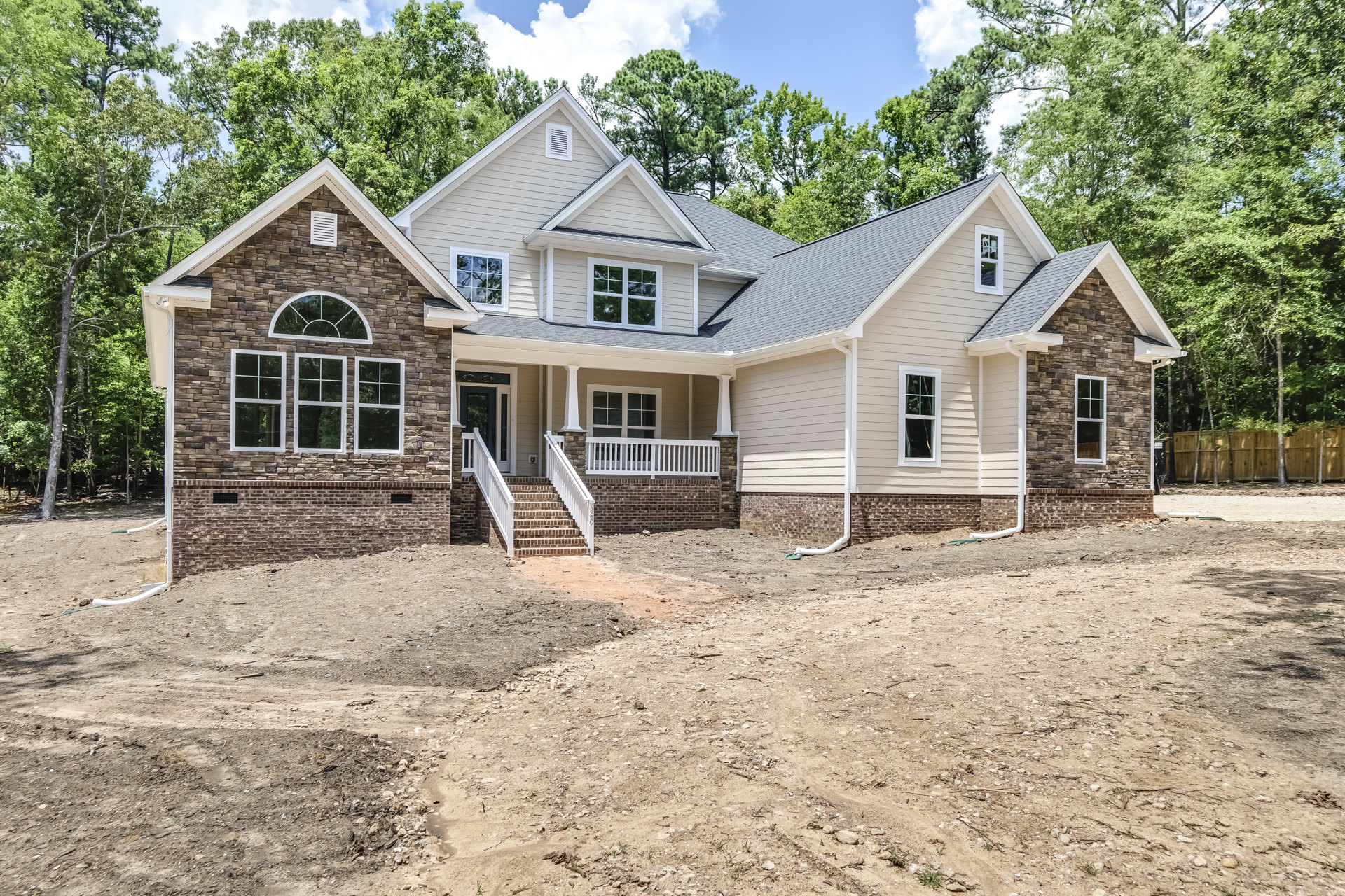 Two-story house with white porch railing, exterior stairs, dirt path and hill in front, broken glass window with white frame, surrounded by trees under cloudy sky