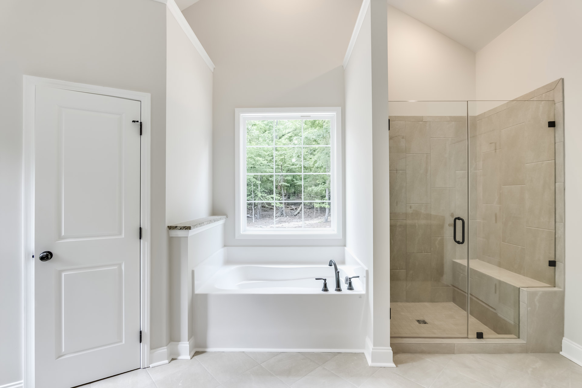 Bathroom with glass shower enclosure and built-in bench, white bathtub with chrome faucets, window overlooking trees, white door with metal doorknob, light-colored tile walls and