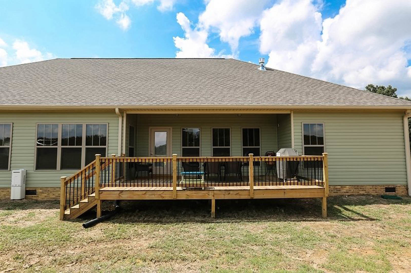 Two-story home with light siding, fenced backyard, wooden deck featuring outdoor chairs, white chimney, large windows, and grassy lawn under partly cloudy sky