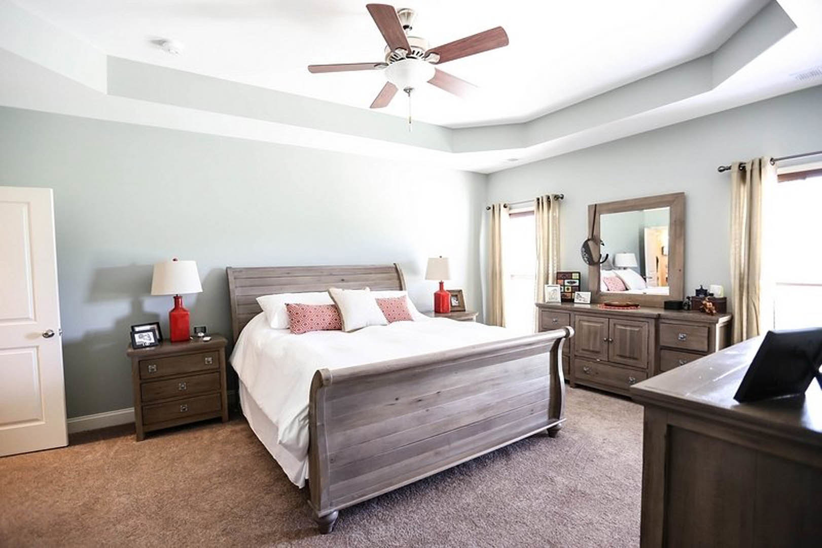 Bedroom with wooden bed frame, white pillows, red lamp with white shade on nightstand, ceiling fan with wood blades, dresser topped with red cup, neutral walls, door partially
