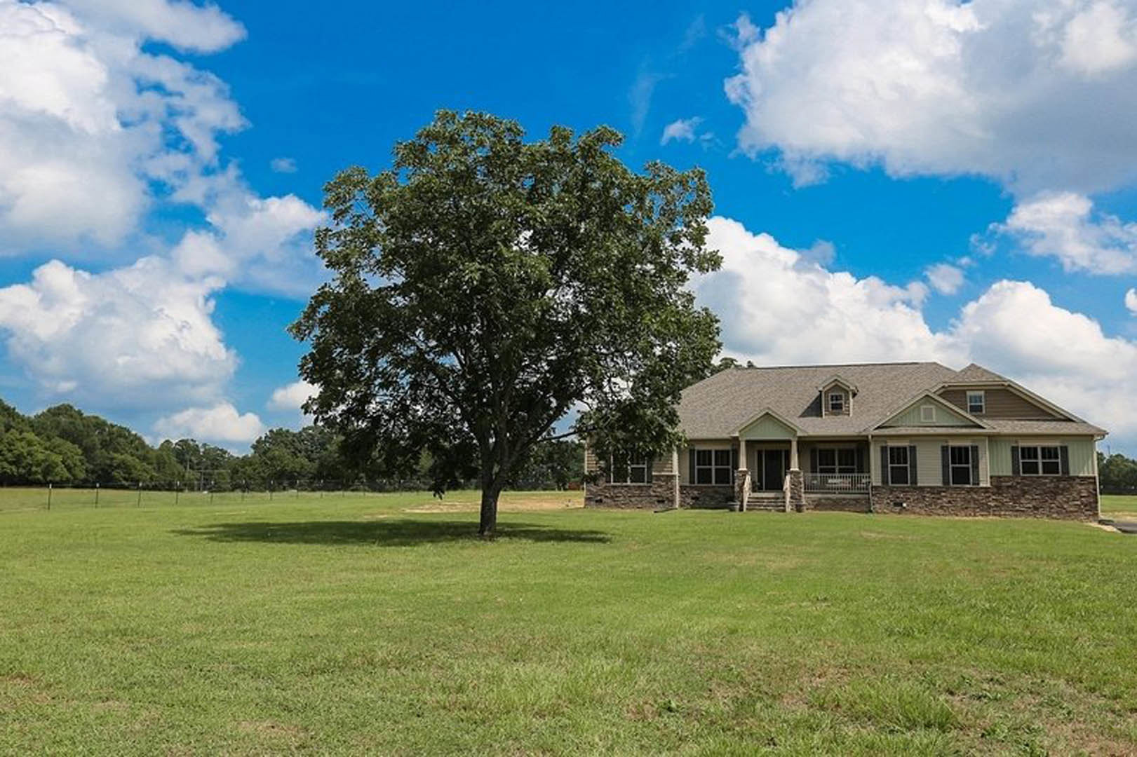 White farmhouse with covered front porch, large windows, and wooden door, set on a grassy field with a mature tree nearby under a partly cloudy sky