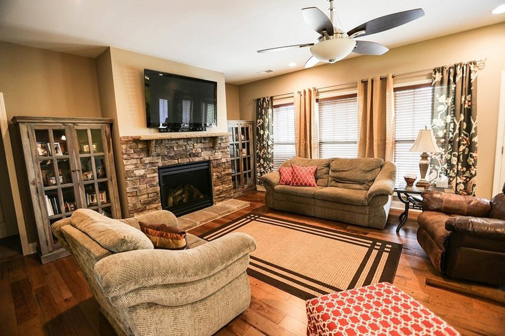 Brown leather couch with patterned pillows faces stone fireplace, wall-mounted TV above mantel, ceiling fan with light fixture overhead, hardwood floors, neutral walls.