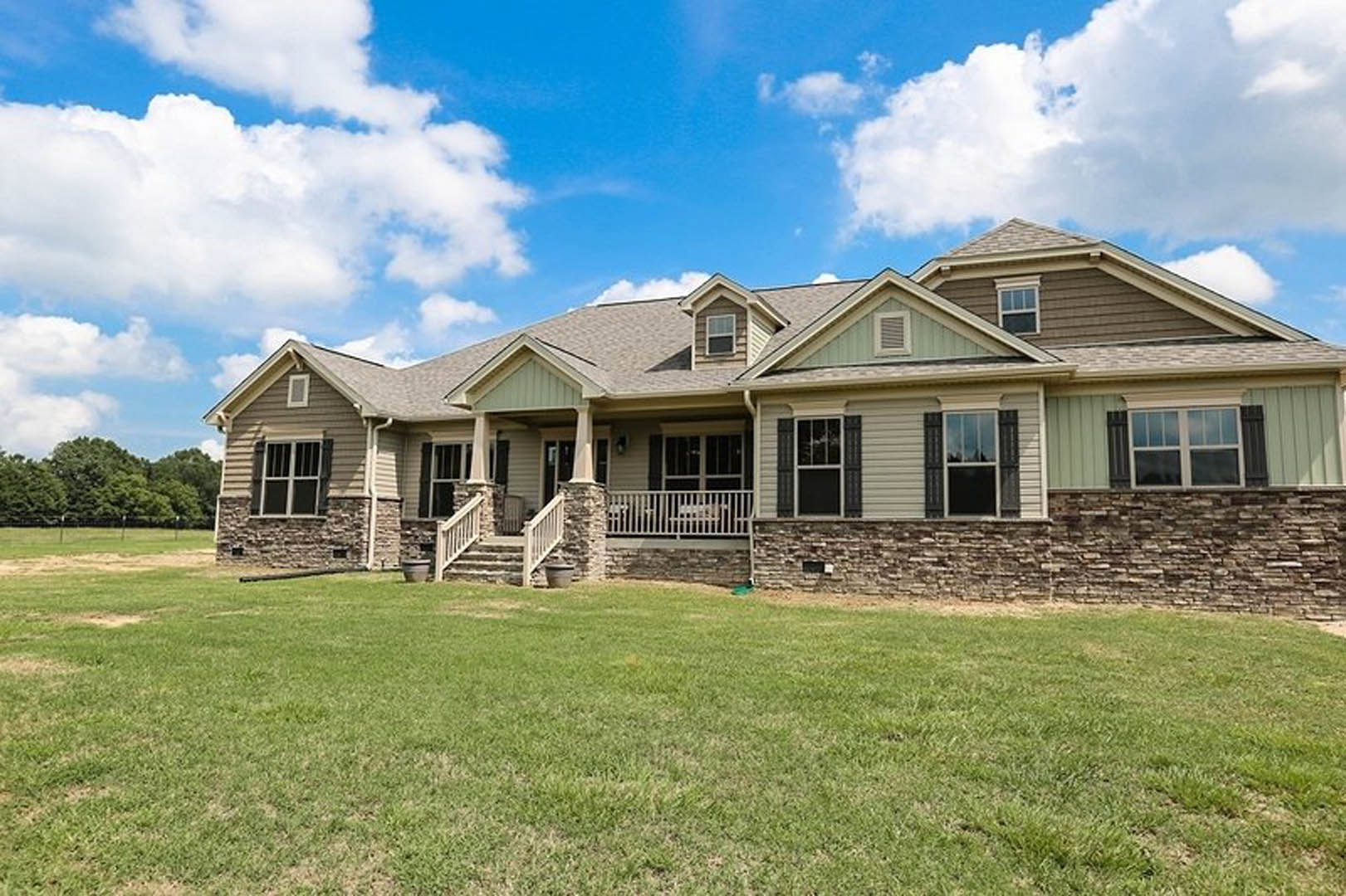 Two-story home with white-trimmed windows, covered porch, stone staircase and retaining wall leading to a green lawn, large tree in background, blue sky with scattered clouds