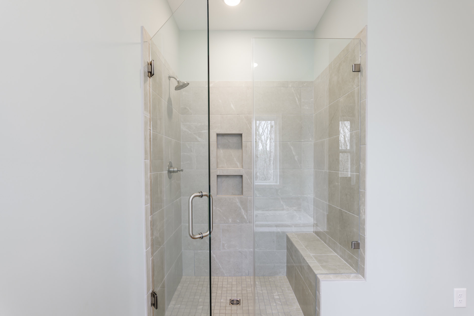 Glass shower enclosure with built-in bench, chrome shower head, light gray tile walls, and tree visible through nearby window