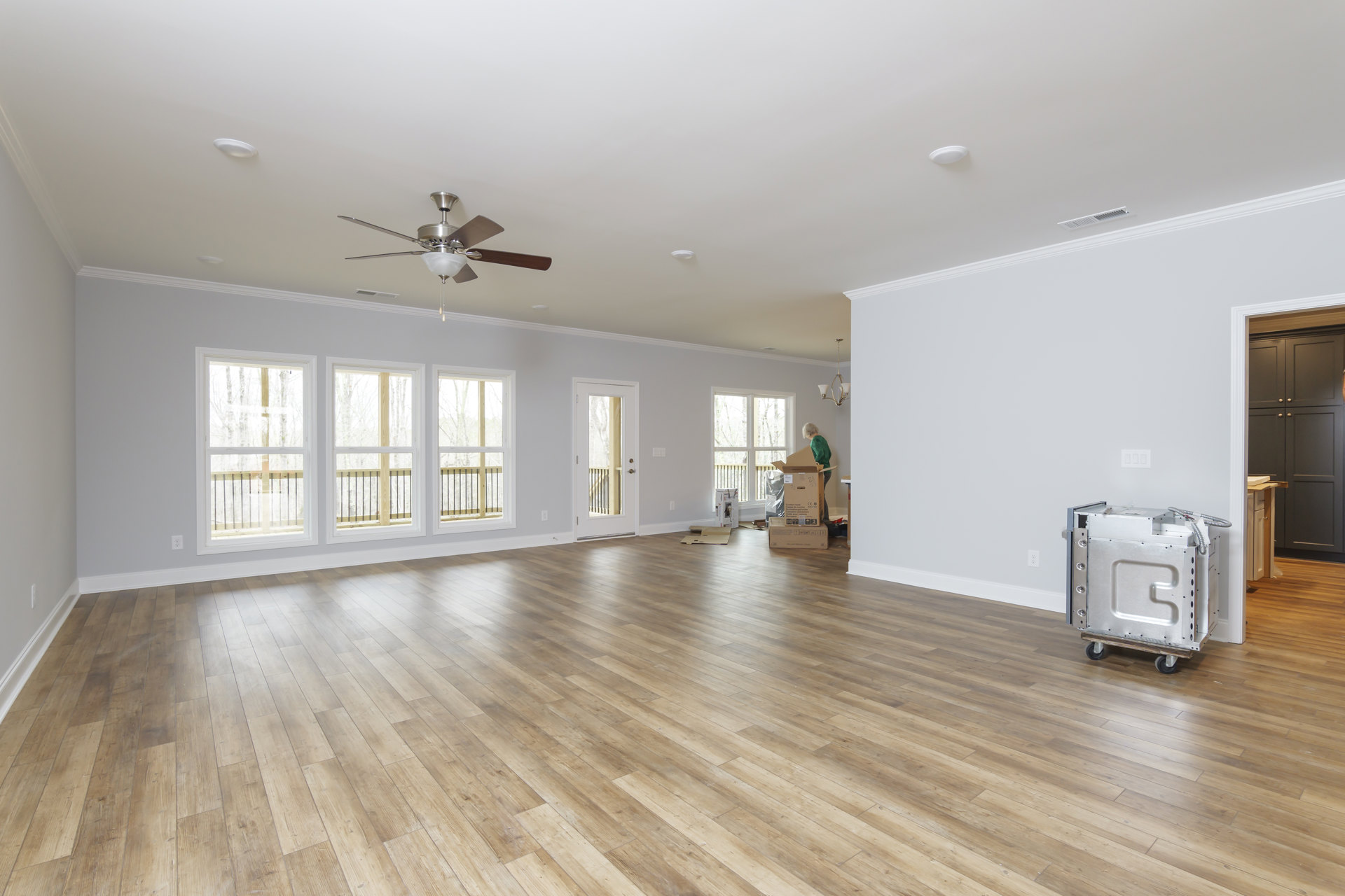 Spacious room featuring hardwood flooring, ceiling fan with light fixture, large windows, and a door with glass panel; metal box on a cart positioned in the center.