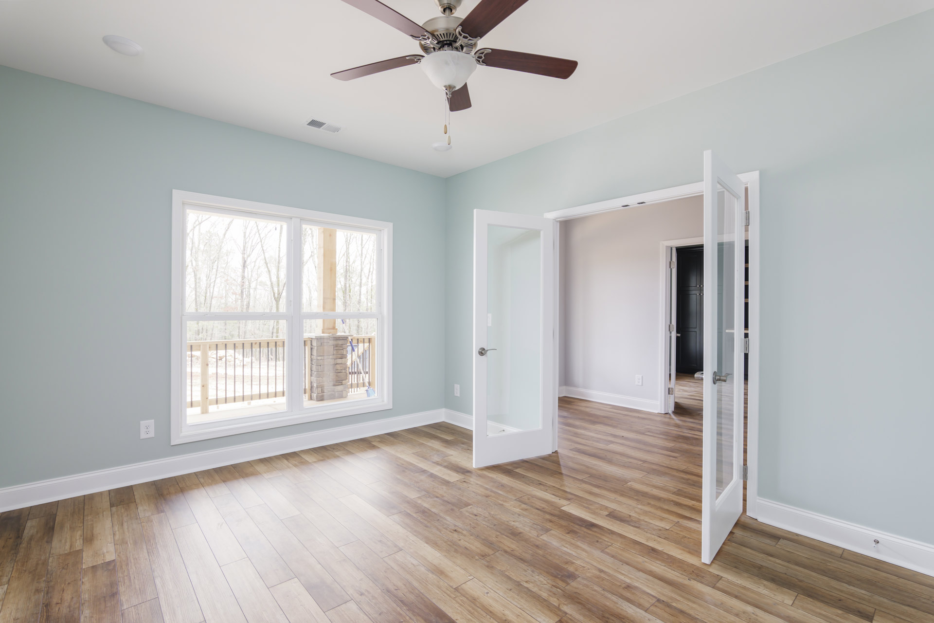 Ceiling fan with light fixture above wood flooring, two doors with silver handles, plaster walls, window beside brick chimney, stone pillar with wood post