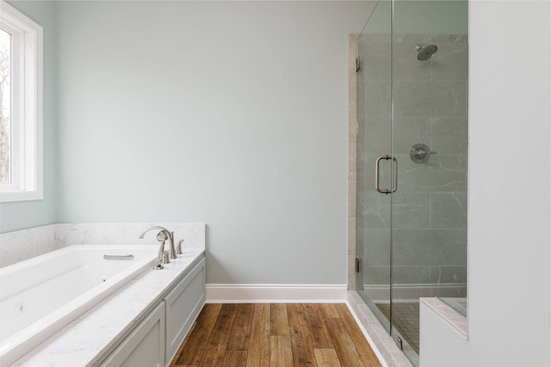 Bathroom featuring glass shower enclosure with chrome handle, freestanding white bathtub, wood flooring, white walls, and window framing view of tree outside