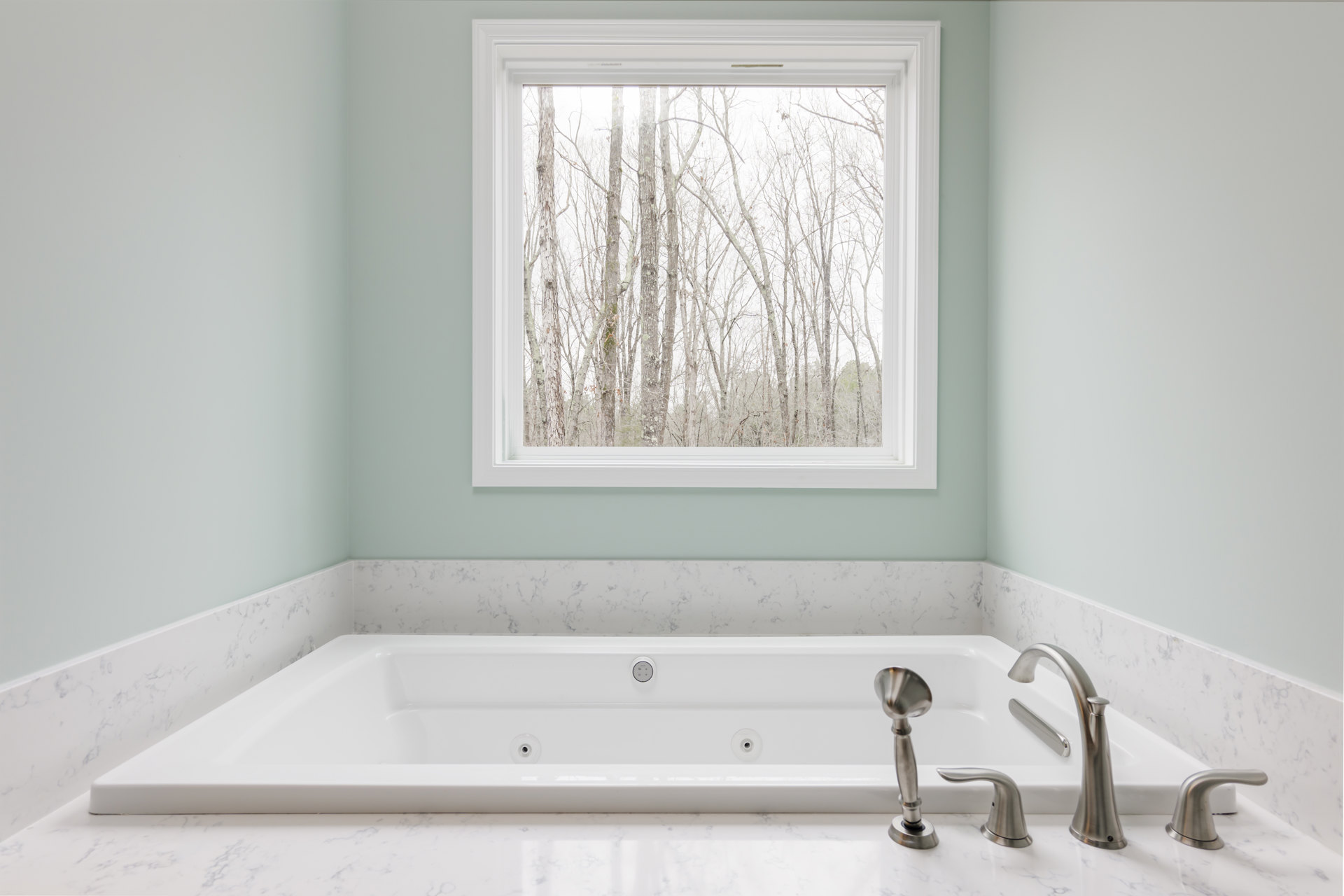 Freestanding white bathtub with chrome faucets beneath a large window, tile wall and trees visible outside