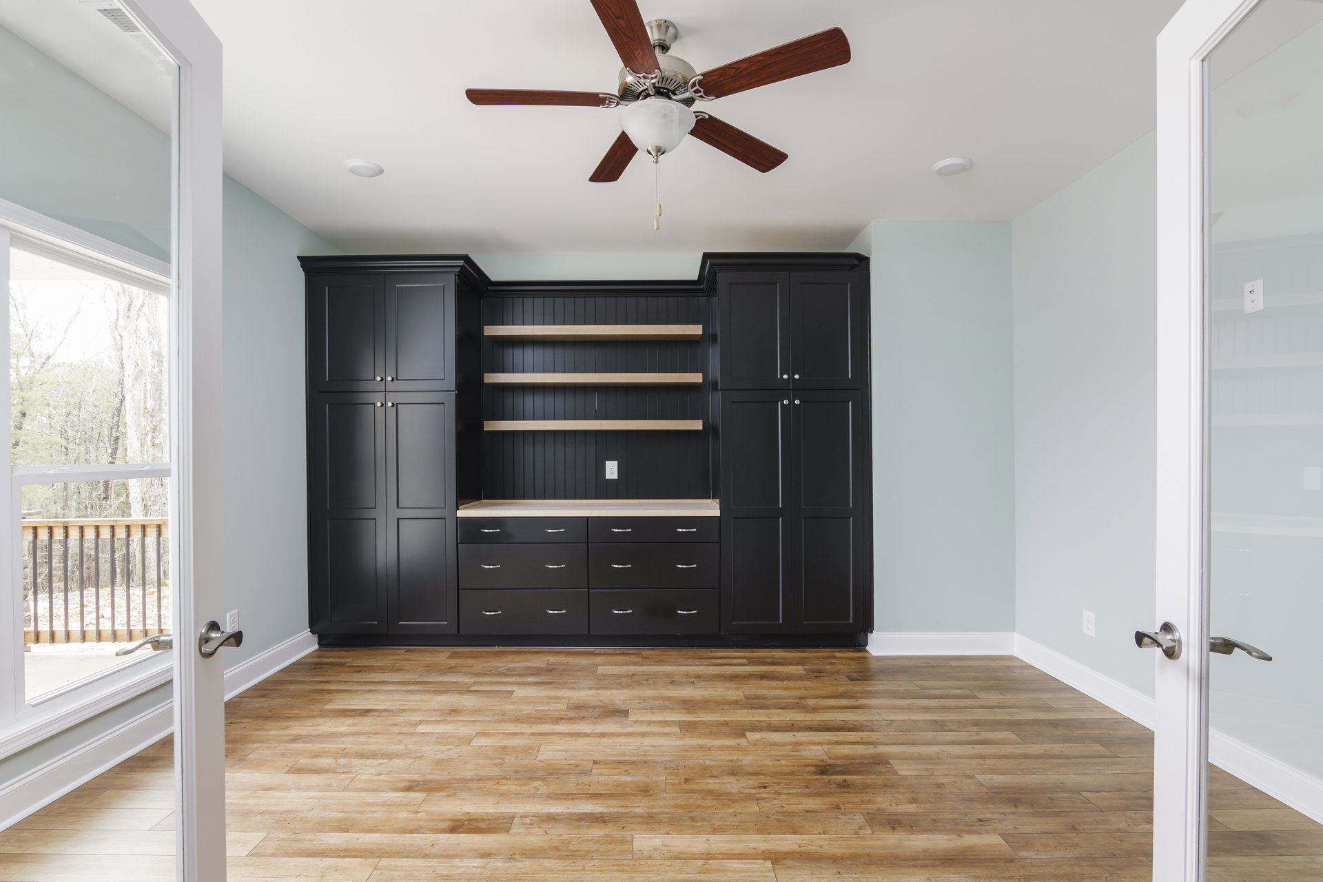 Ceiling fan with light fixture above wood flooring, black cabinet with open shelves, wood railing and post, neutral walls