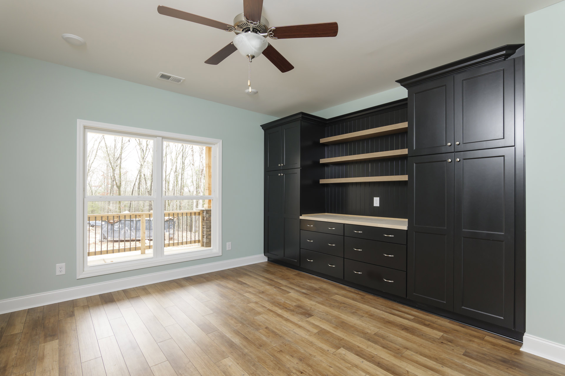 Black cabinets with silver knobs, wood flooring, ceiling fan with light fixture, window showing exterior railing, and built-in drawers along the wall.