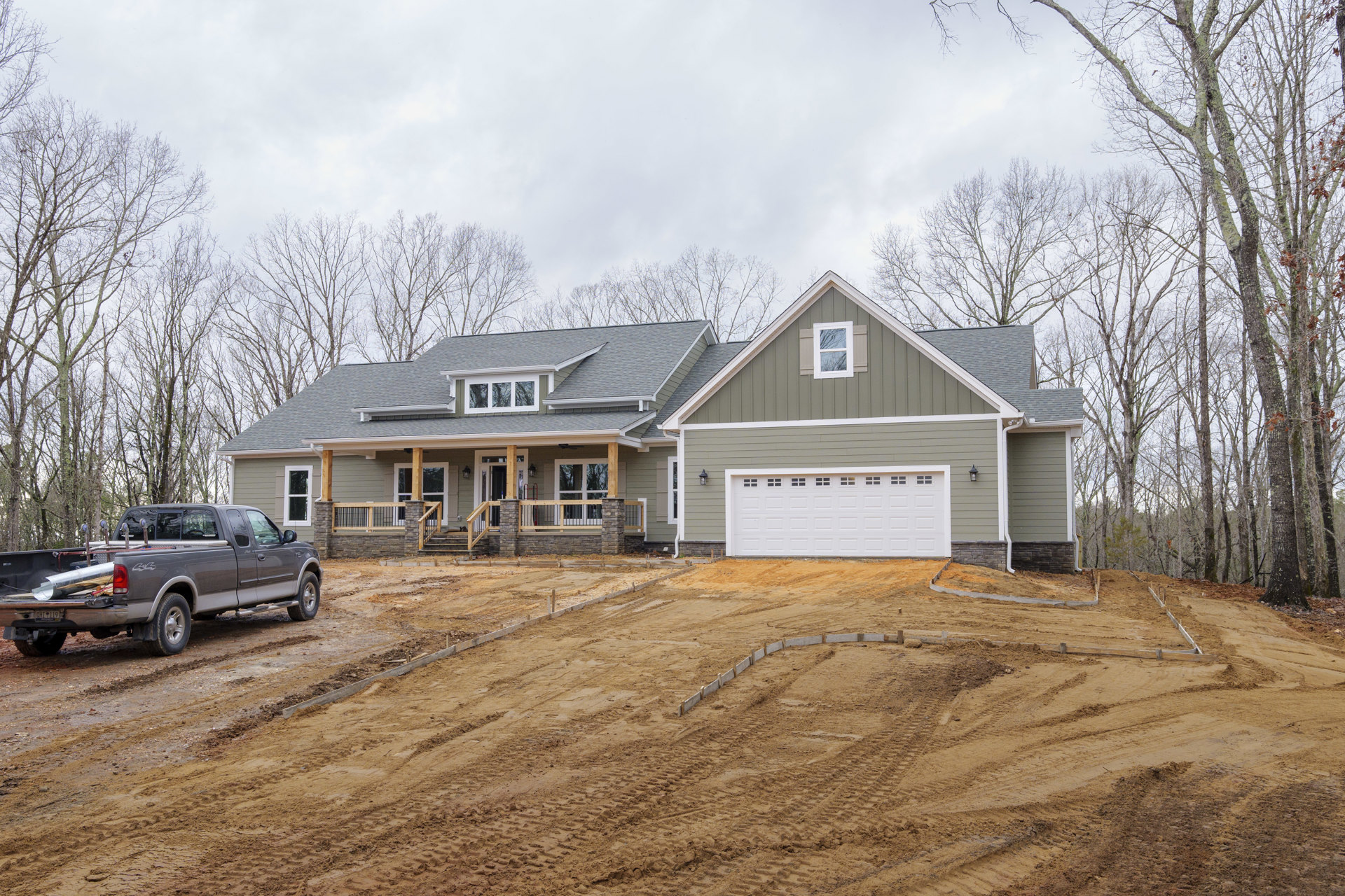 White garage door with upper windows, white-framed window, dirt driveway with tire tracks, pickup truck carrying a pipe parked in front, cloudy sky above, trees in background