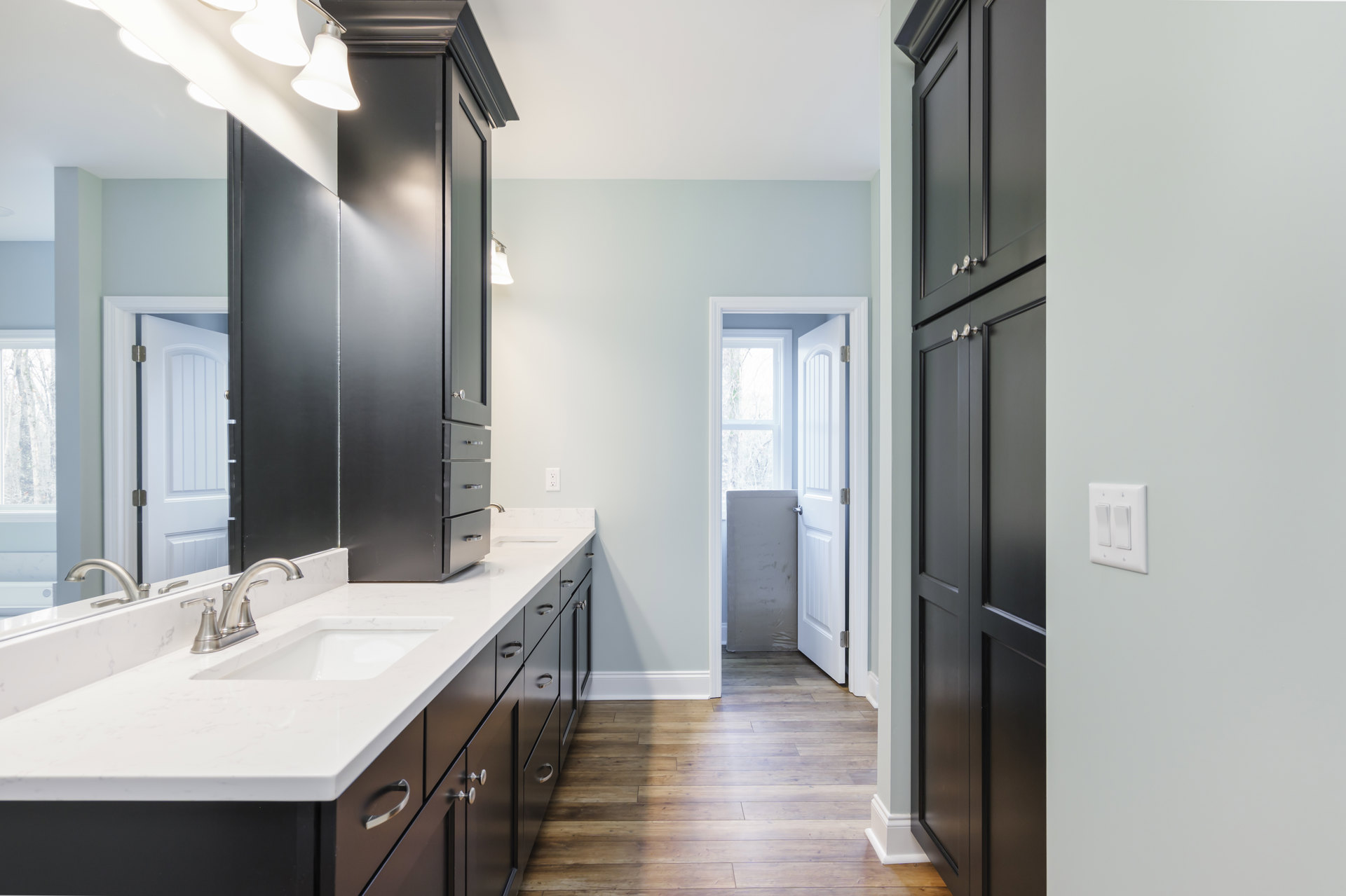 Bathroom featuring black cabinetry with white countertops, undermount sink, chrome faucet, white walls, white door partially open to a window, and close-up views of a cabinet