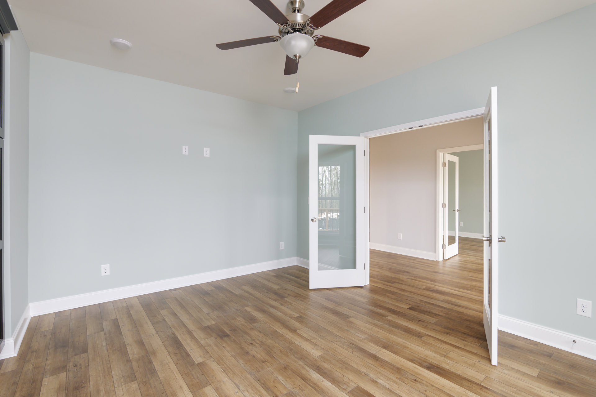 Wood flooring and white walls in a bright room with a ceiling fan and light fixture, glass-paneled door, and white trim.
