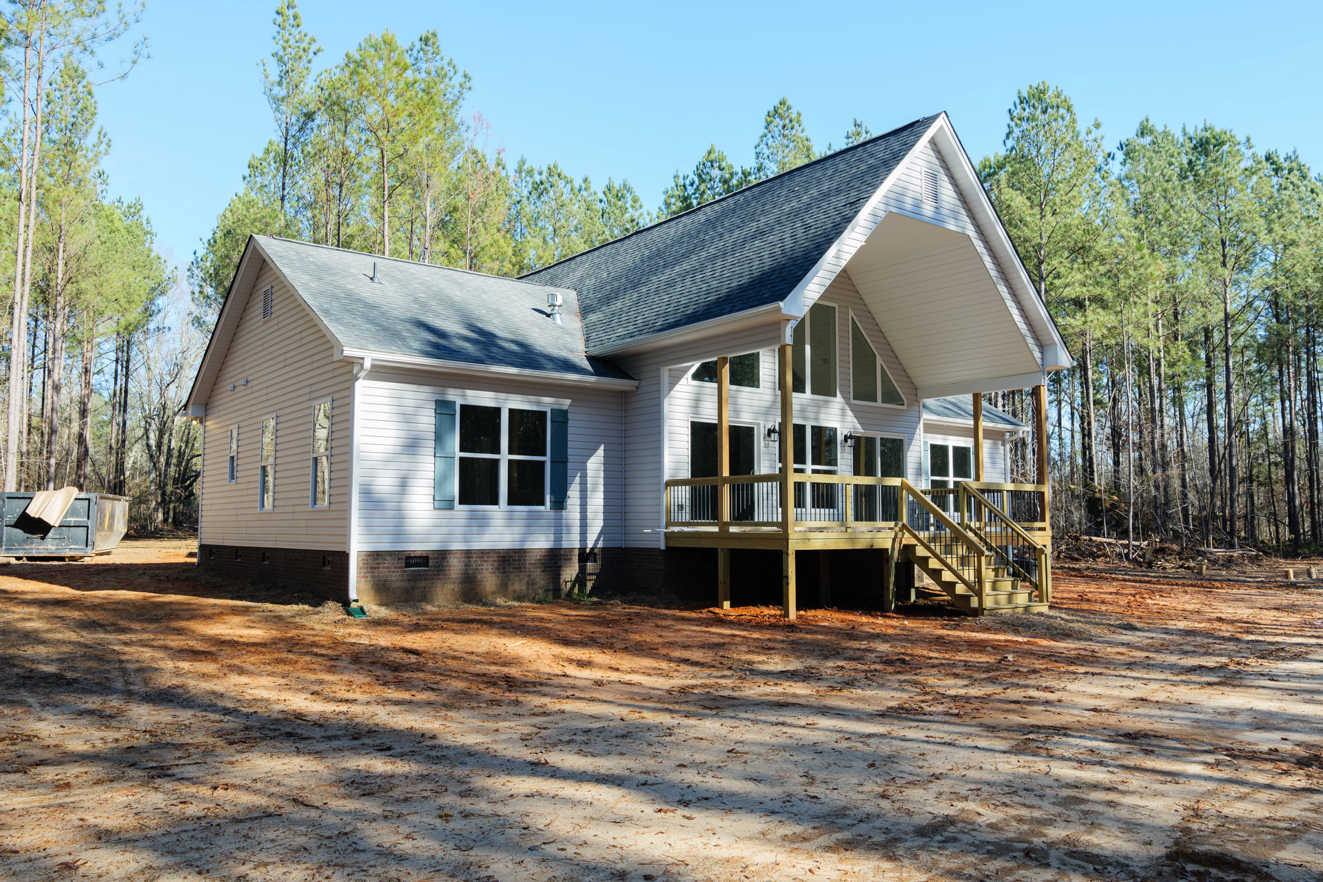 Two-story house with white-trimmed windows, wooden porch and deck with railing, dirt path covered in leaves, trees in background, metal container near entry.