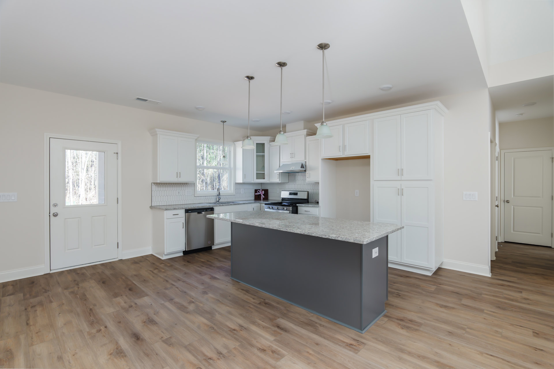 Kitchen with wood flooring, central island featuring built-in stove and oven, white cabinetry, stainless steel sink, white door with window and silver hardware, large window