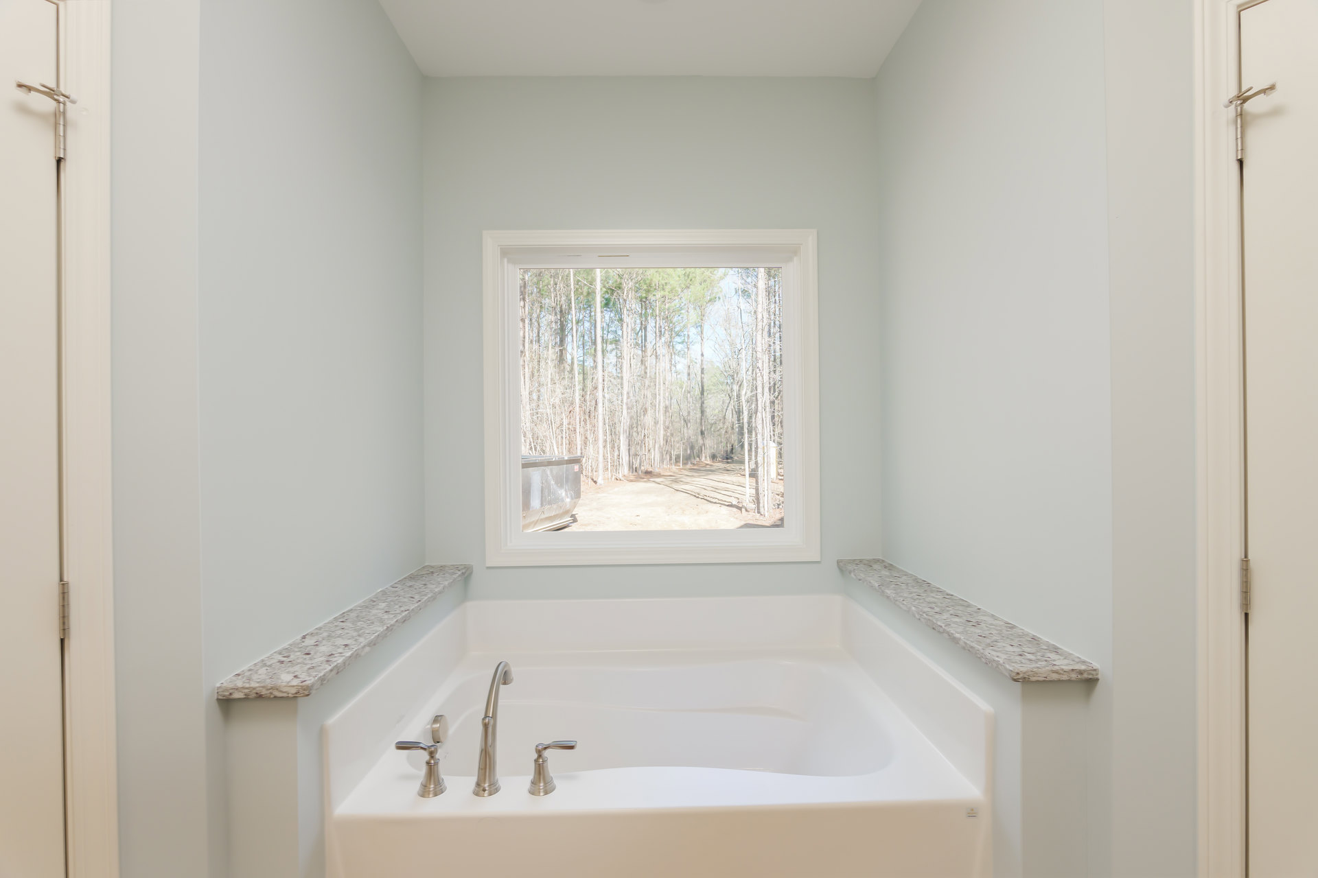 Freestanding white bathtub beneath large window with tree views, chrome faucet, light gray tile walls, and minimalist bathroom decor
