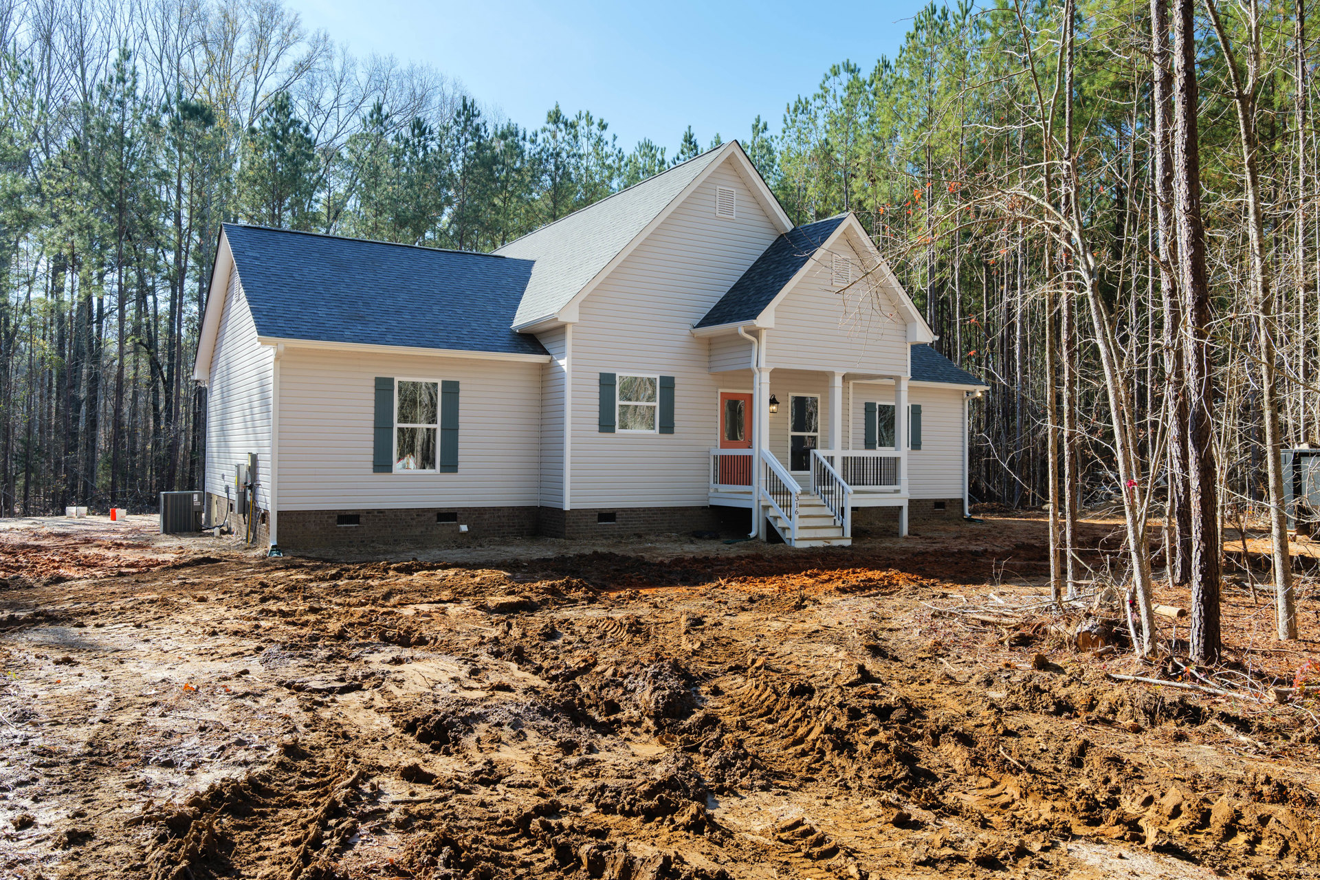 Partially built house with exposed framing, white window frames, and unfinished porch, surrounded by dirt lot and mature trees in the background