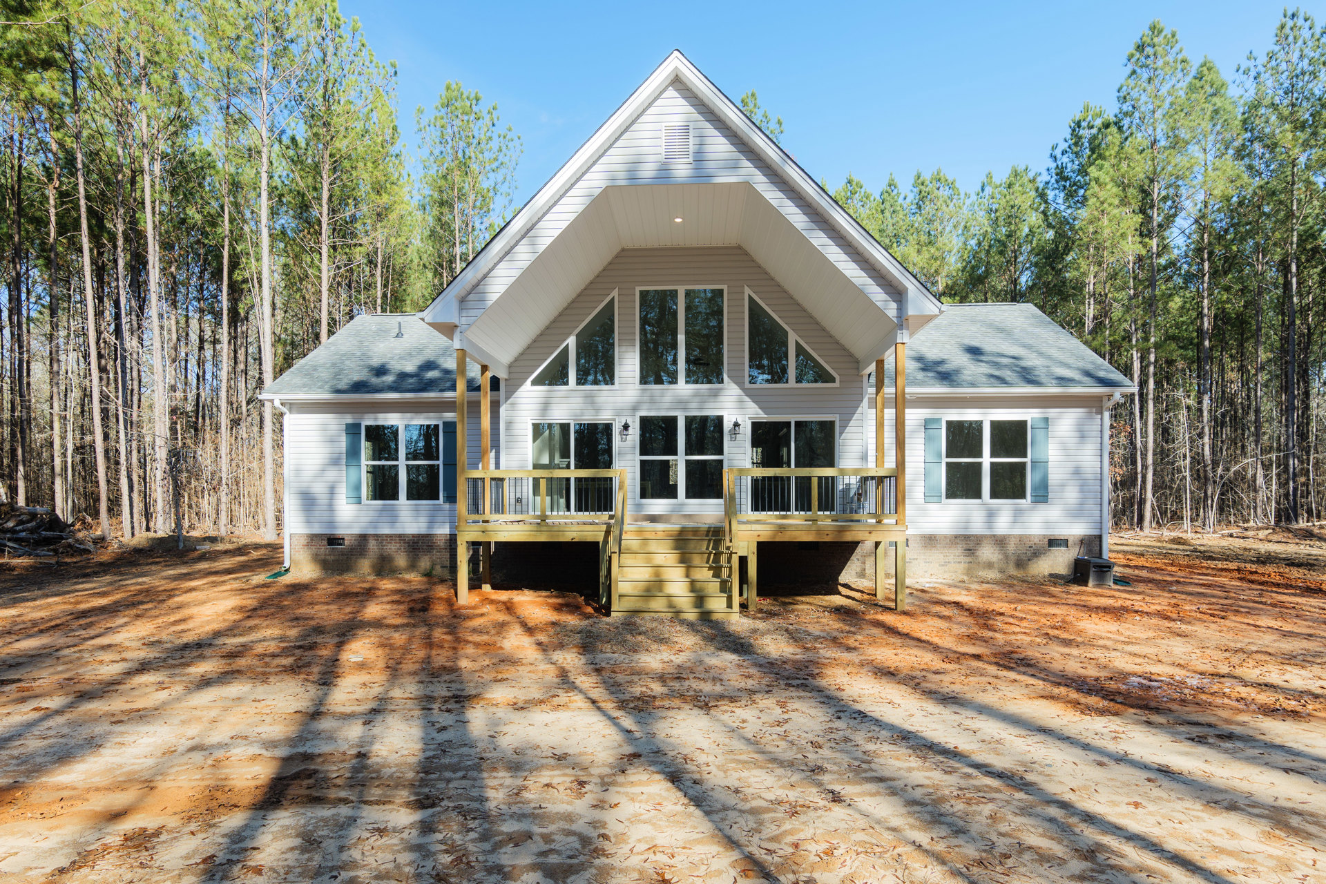 Wooden porch and deck with stairs leading to a grassy yard, mature trees in the background, white-framed windows on light-colored siding