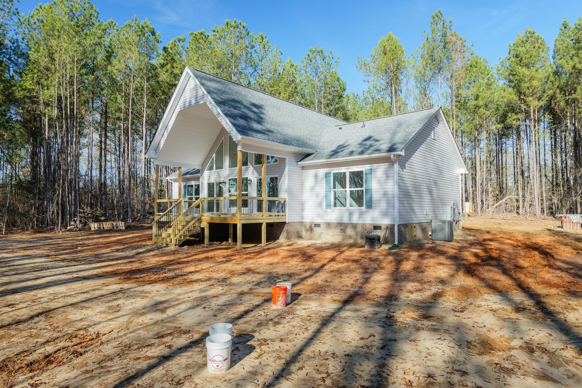 Wooden deck with railing surrounded by trees, buckets of paint on the ground near white cottage-style home, porch with windows and door visible