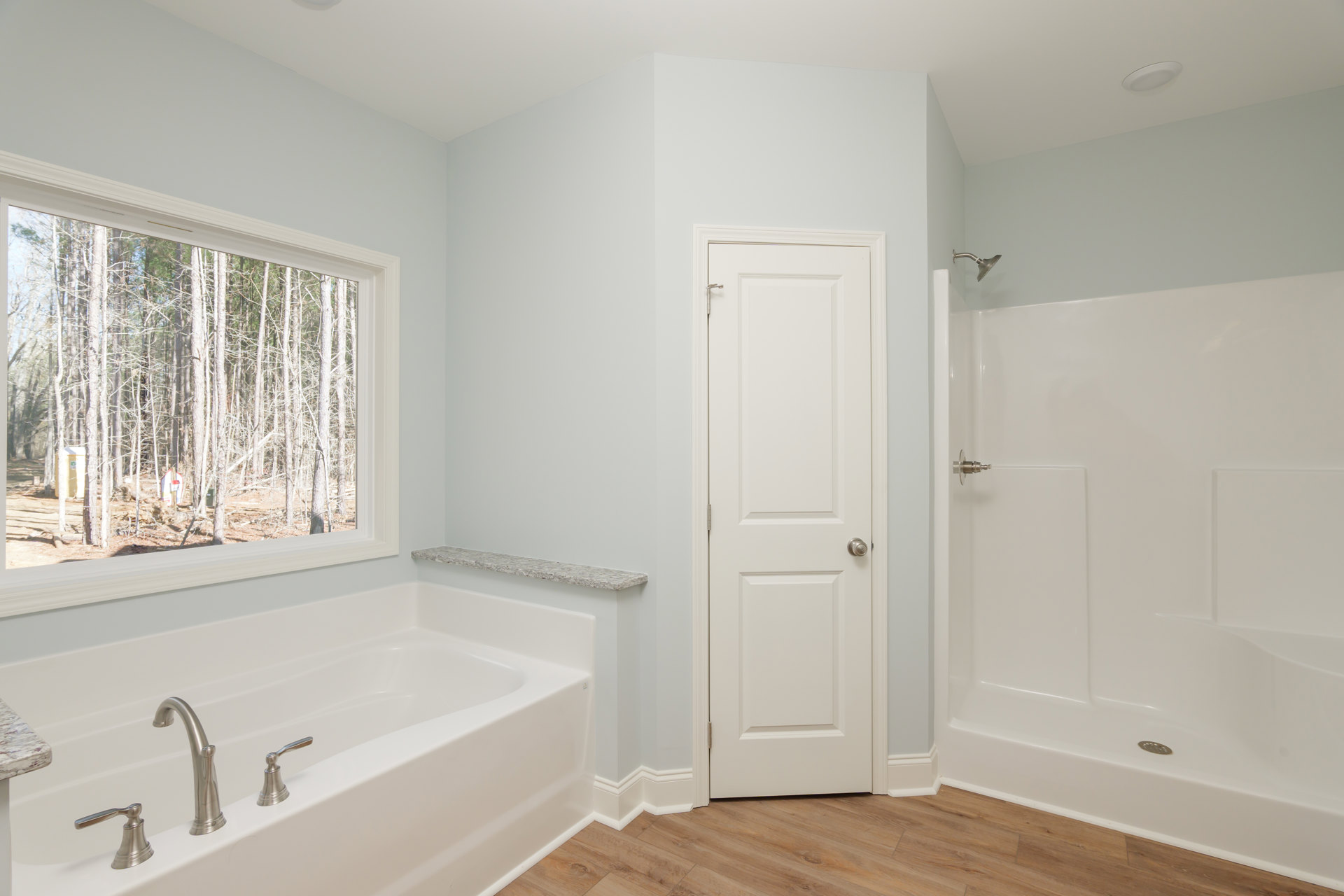 Bathroom with white tile walls, freestanding bathtub, glass shower enclosure, silver fixtures, white door with silver knob, window overlooking trees.