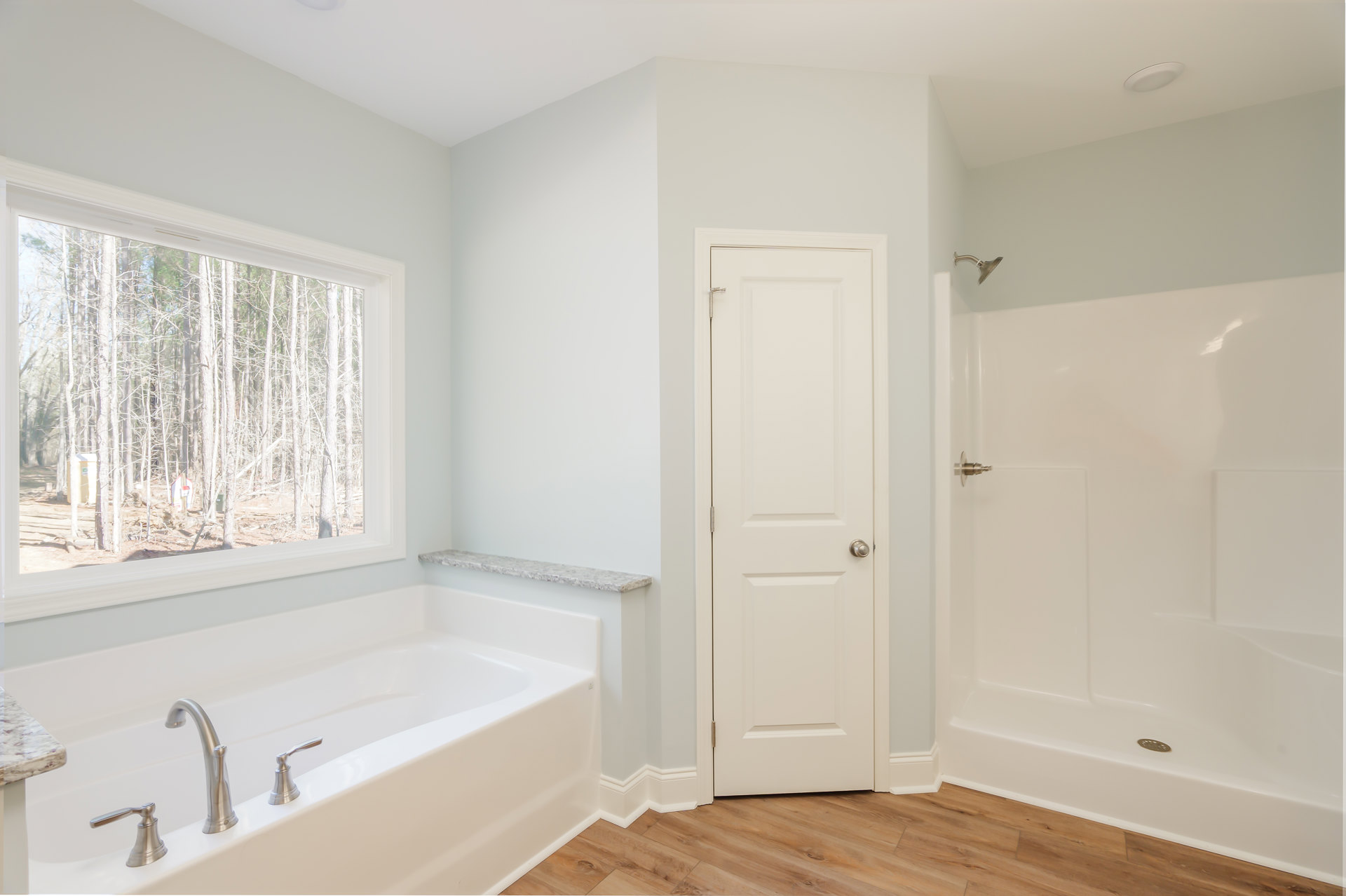Bathroom with freestanding bathtub beneath a window overlooking trees, white tile walls, silver faucet, white shower with drain, white door with silver knob, and close-up of stone