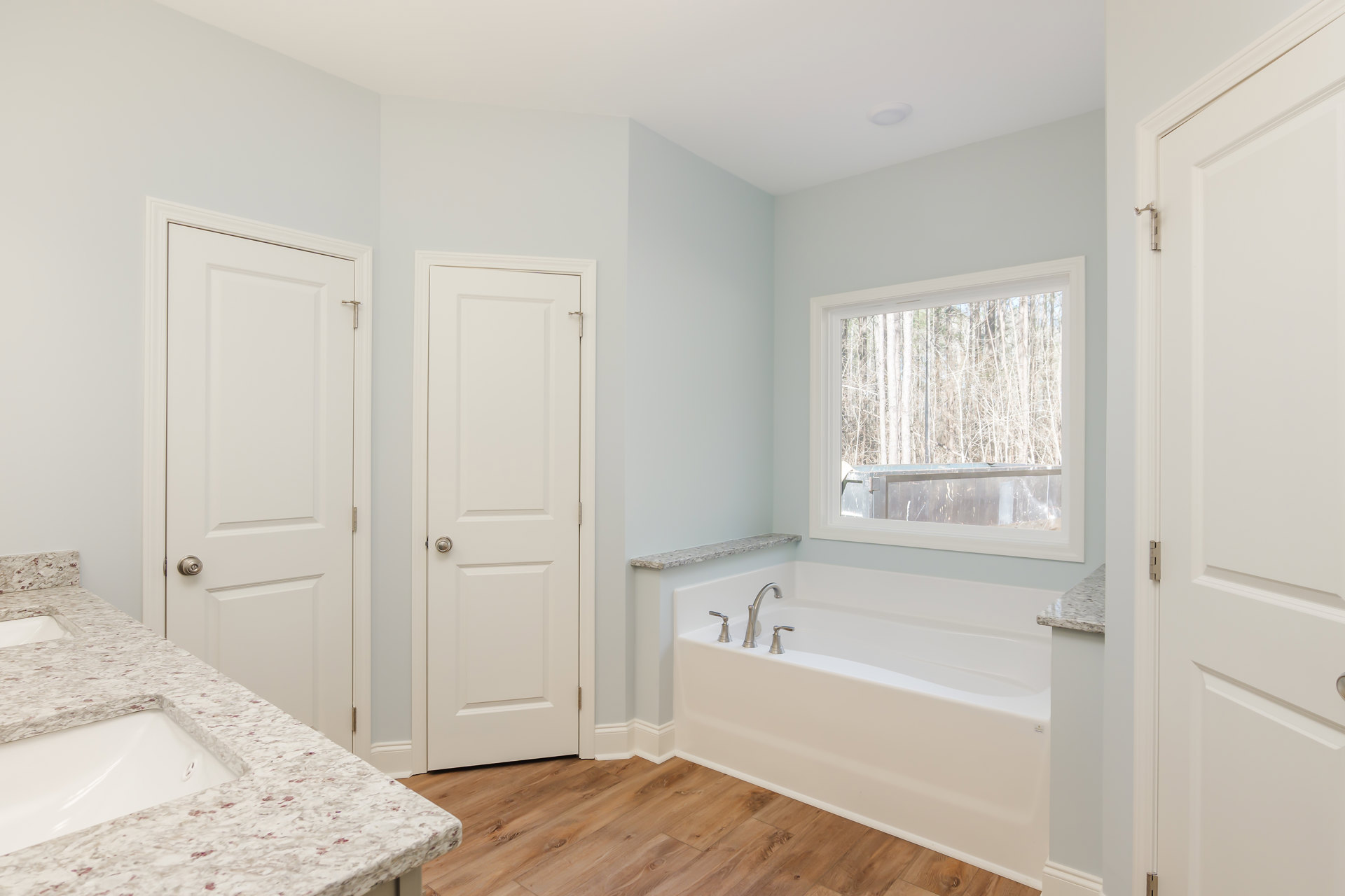 Bathroom featuring a freestanding bathtub with chrome faucets, marble countertop vanity, large wall mirror, white cabinetry, and a window overlooking trees.