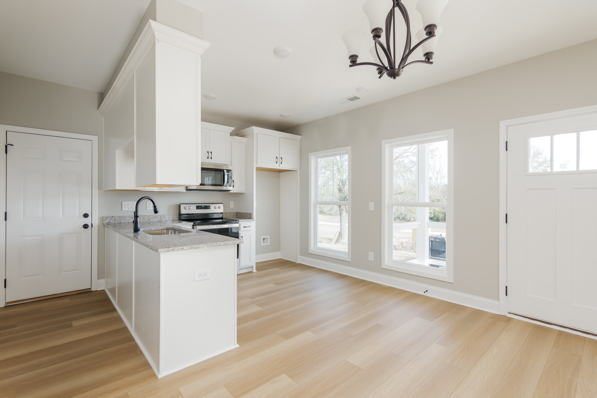 Kitchen with wood flooring, white cabinetry, white island with built-in sink, black hardware on doors, large window showing tree outside, chandelier hanging above island, broken