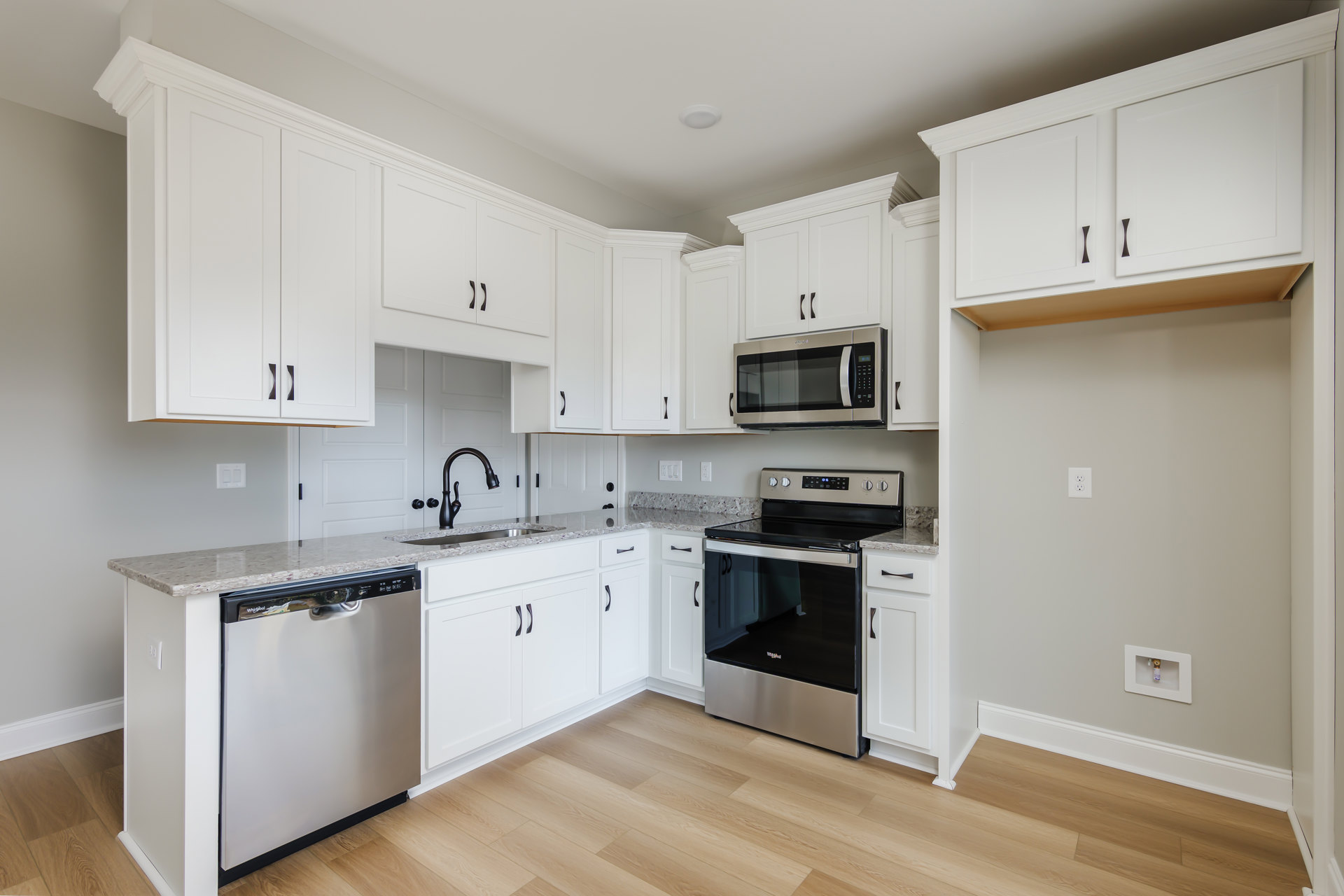 White kitchen cabinets, black and silver oven, microwave with door, silver dishwasher with black handle, close-up of chrome faucet, light countertops, hardwood flooring