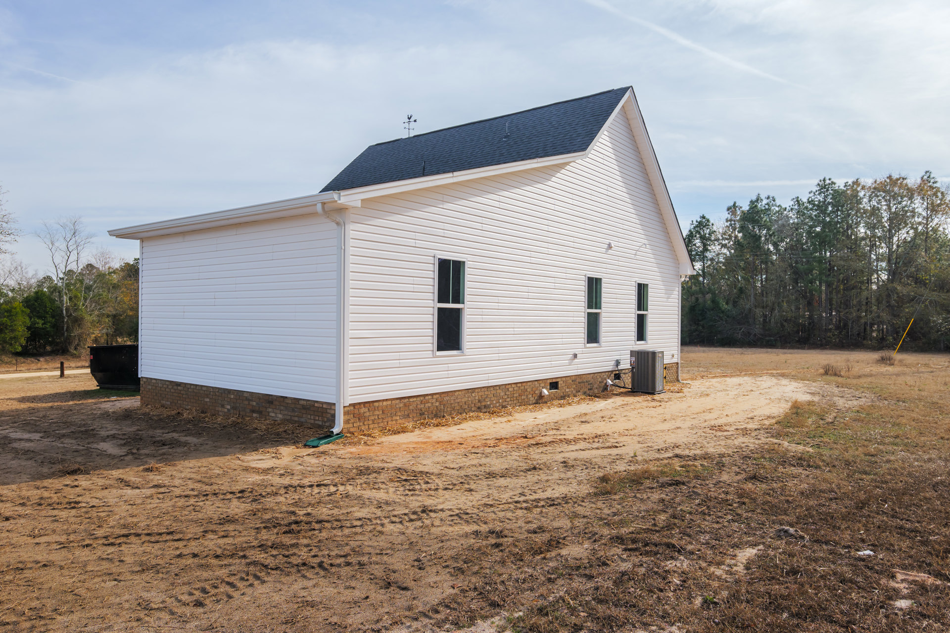 White siding house with black shingle roof, white-framed window, dirt yard, brick wall, large gray HVAC unit, surrounding trees and overhead power lines