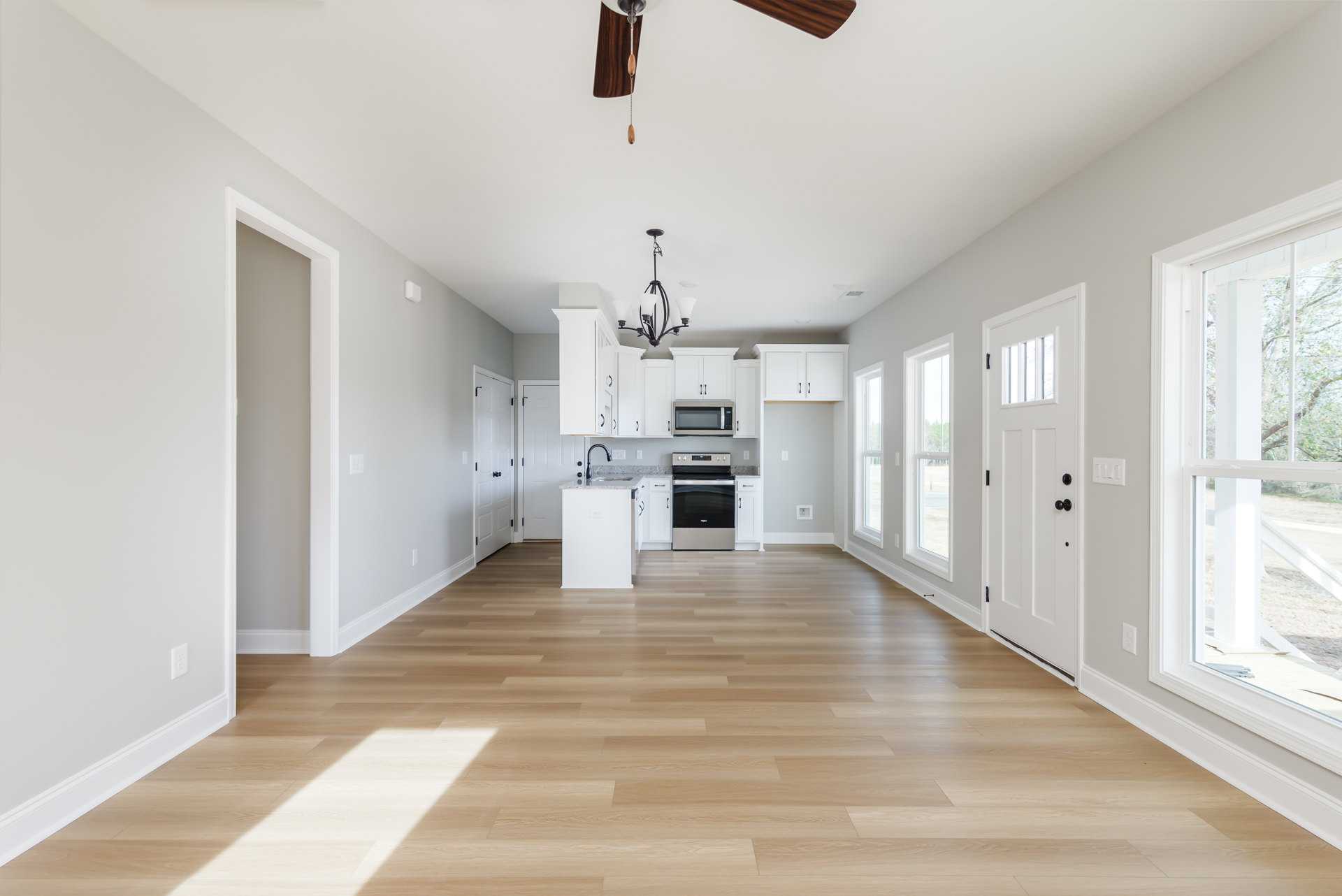 Open-concept kitchen with white cabinetry, stainless steel stove and microwave, hardwood flooring, white column, ceiling fan, and window overlooking a leafy tree.