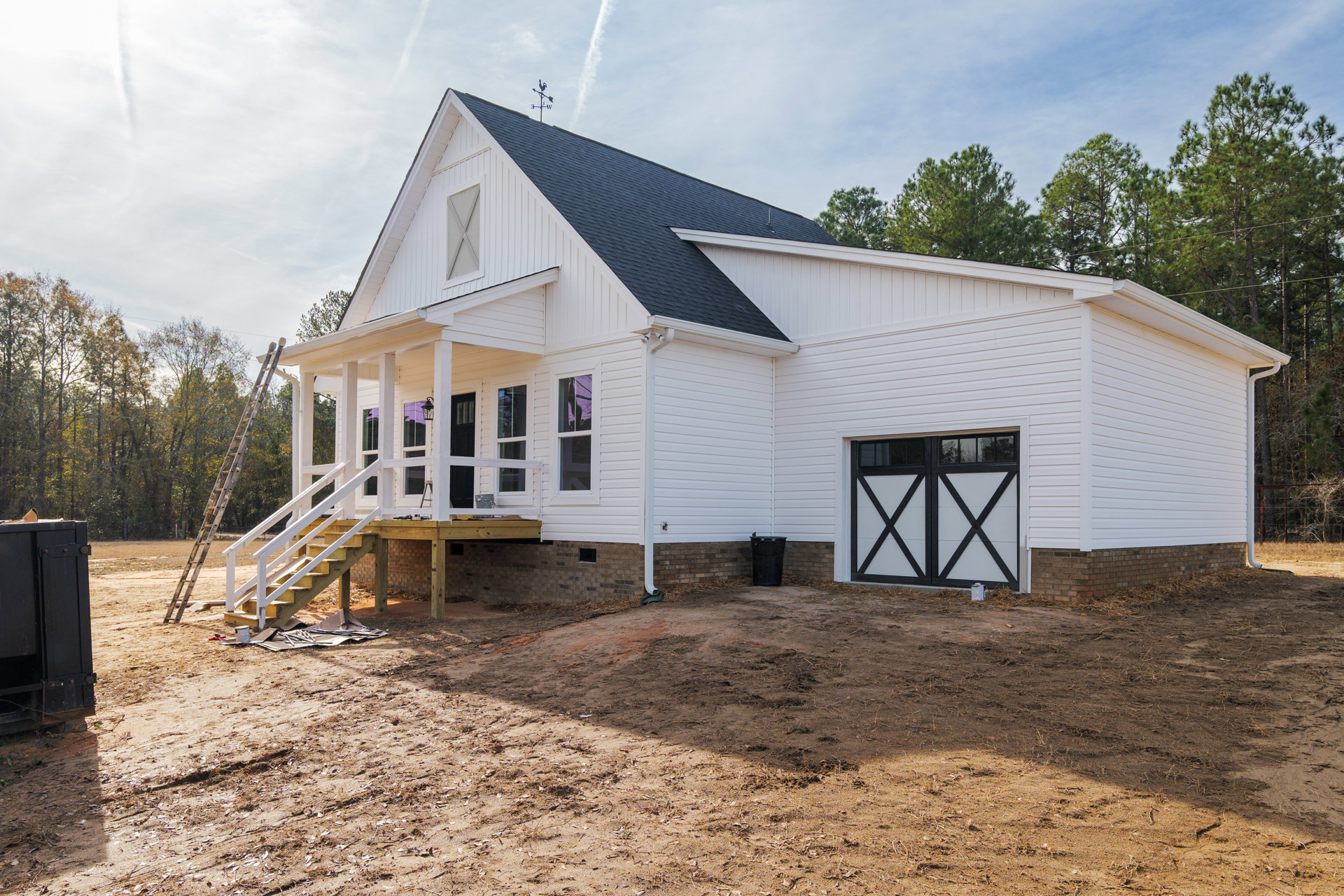 White siding house with black trim, attached garage with white door, covered front porch, dirt yard with trash can, ladder leaning against exterior wall, cloudy sky overhead.