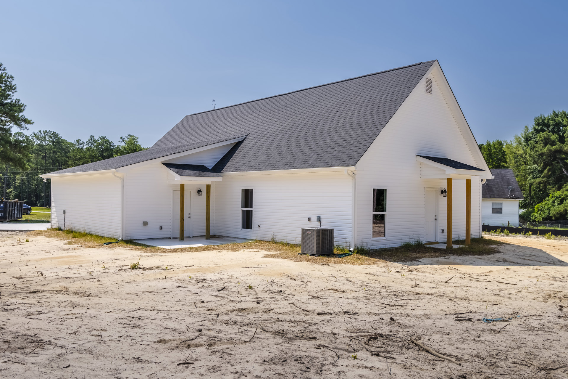 White siding house with black shingle roof, grey utility box on dirt ground, large metal dumpster with logo, trees in background, clear sky above.