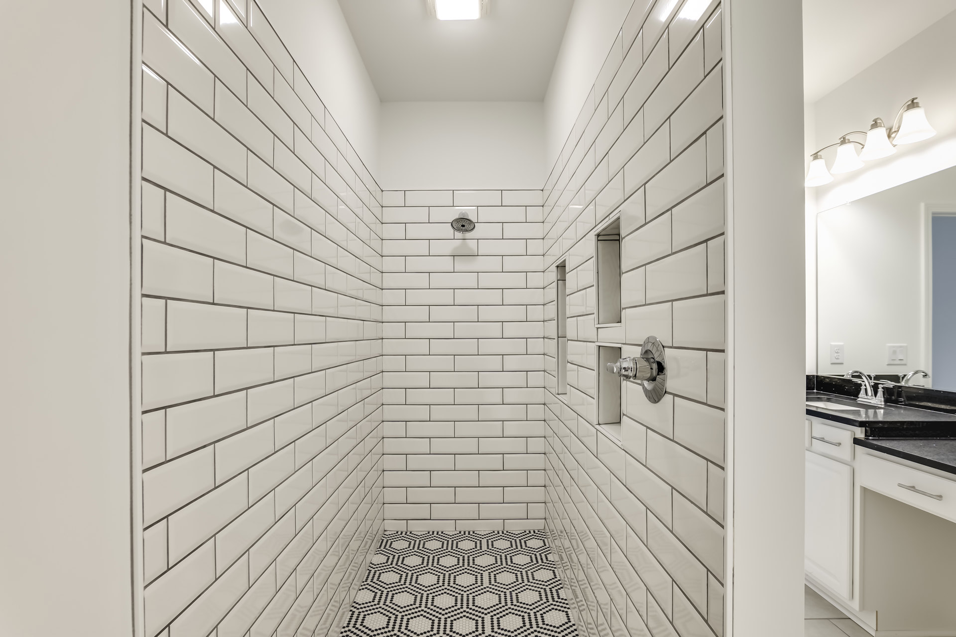 White tiled shower walls with a black and white patterned tile floor, silver shower fixtures, and a white-shaded light fixture
