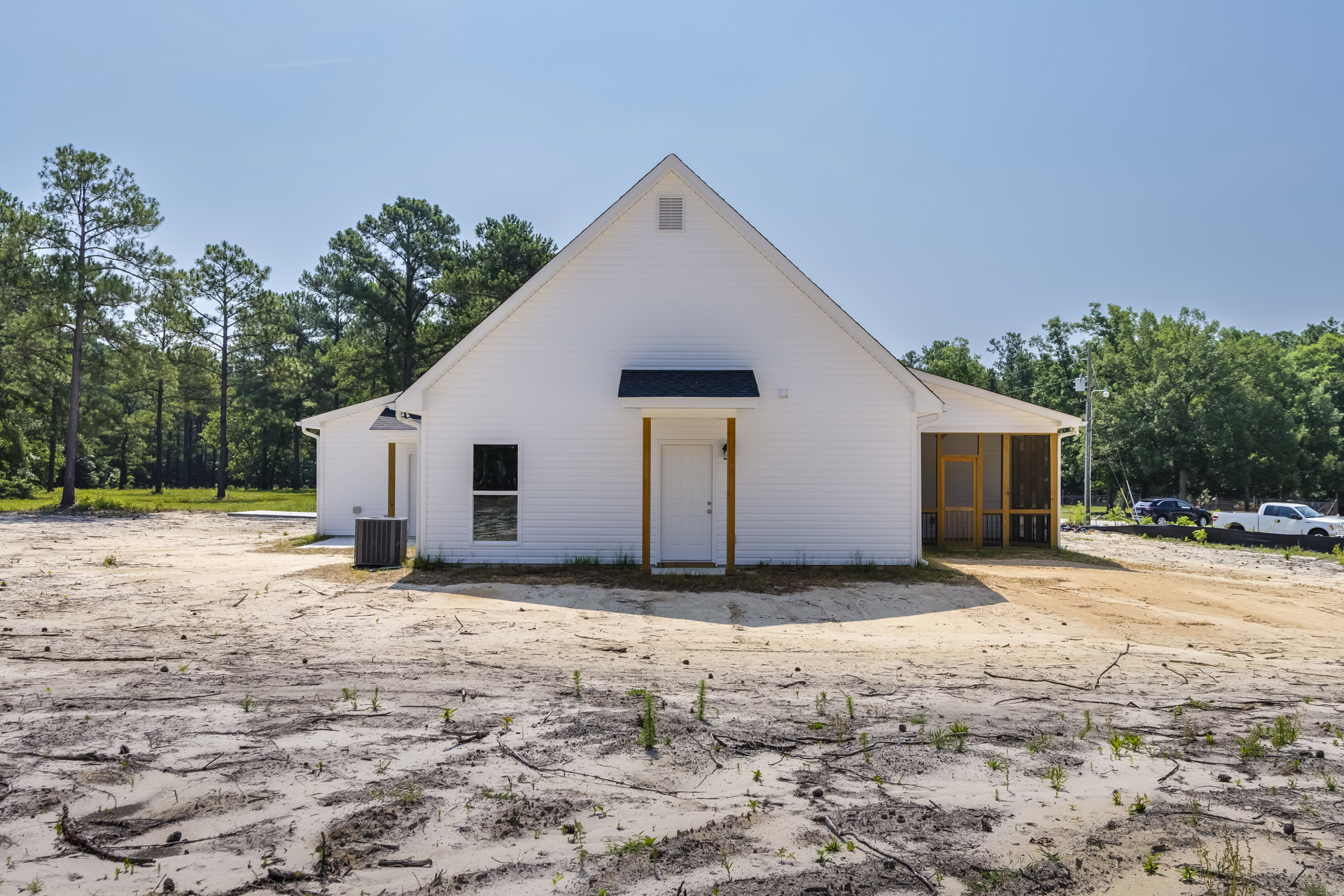 White house with black roof, white door, and white-framed window; large metal box on dirt area in front; trees in background.